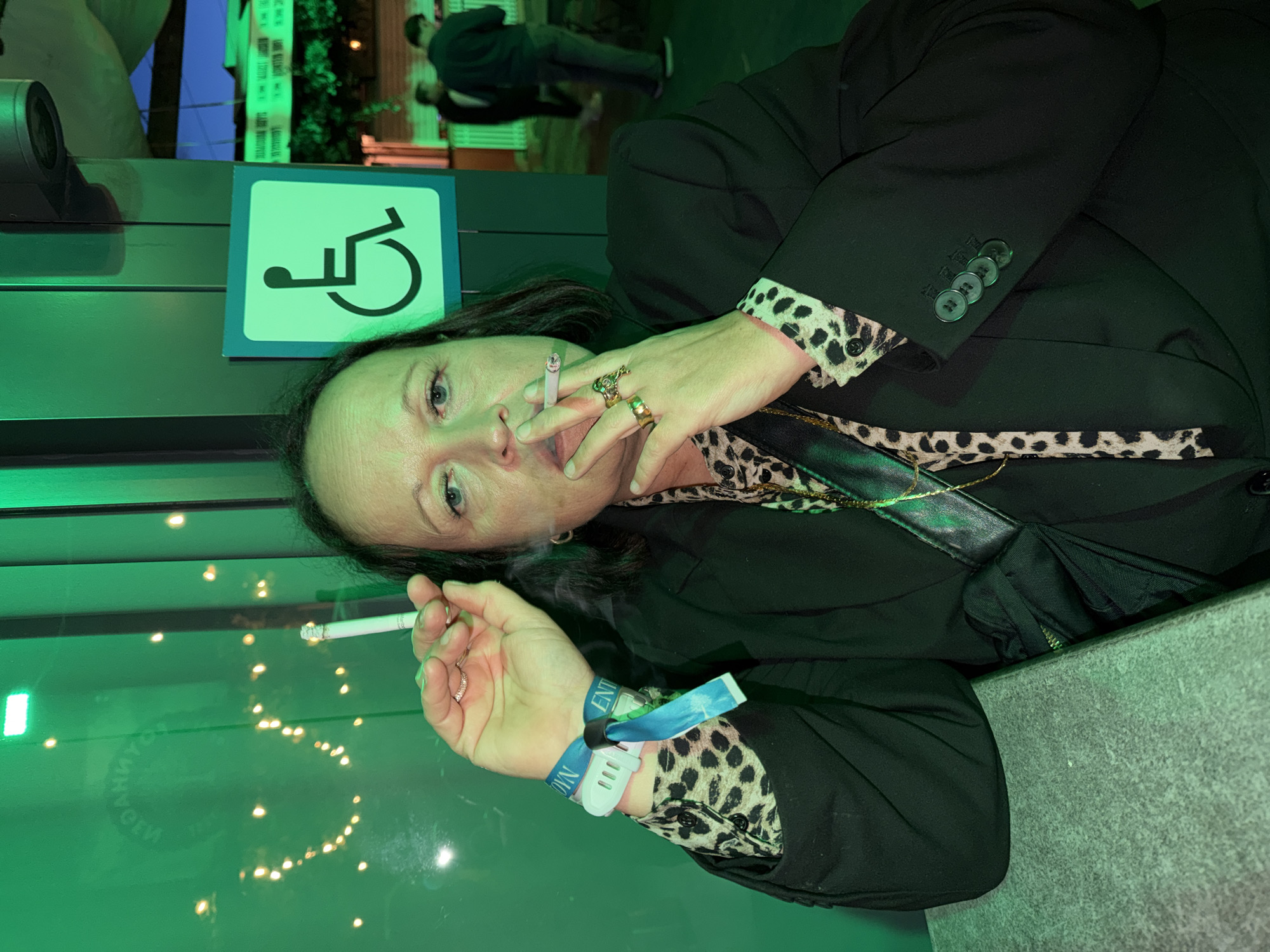 The image captures a middle-aged individual, likely female, smoking two cigarettes in a public or semi-public outdoor setting in Tønsberg, Norway, under dominant green artificial light. The subject looks directly forward, holding one cigarette between her lips, from which smoke rises, and another between the index and middle fingers of her left hand. She wears a black blazer over a leopard print collared shirt. Her left wrist displays a white smartwatch-like device with a blue fabric band and an additional blue fabric wristband, both potentially event passes. Multiple rings adorn her right hand, including a distinctive gold-toned ring with a green, possibly animal-head, design on her middle finger. A thin gold chain necklace is visible. The subject leans on a light-grey, textured surface. The background on the left shows a reflective green surface with indistinct reflections of string lights and mirrored text. On the right, a light-green square sign with a black International Symbol of Access is affixed to a dark green structure, above which a security camera is mounted. Beyond this, two blurred male figures are visible in a further background, suggesting an open-air area under a dark sky.