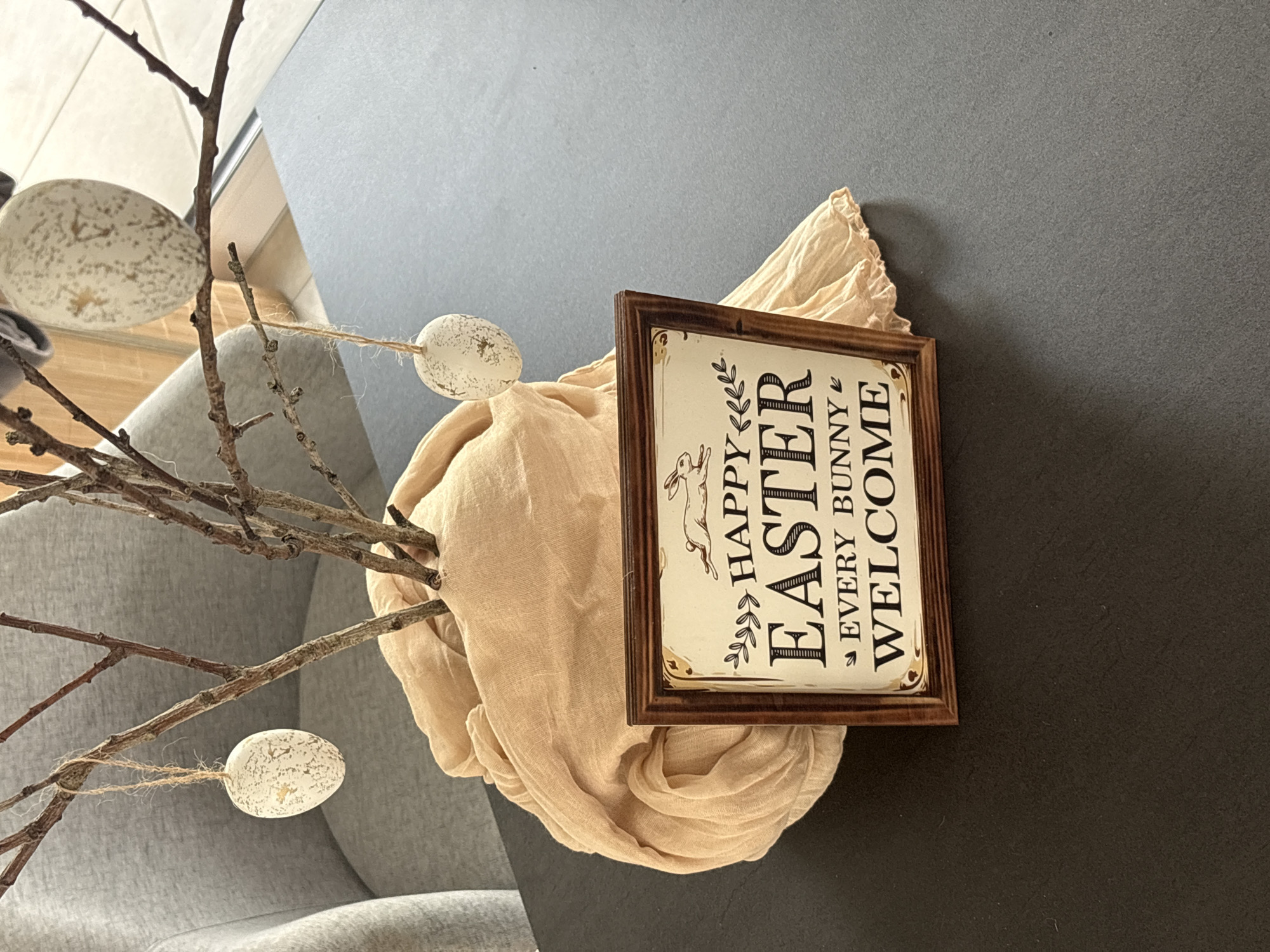 A still-life photograph depicts a decorative Easter arrangement. Several bare branches, adorned with three speckled, egg-shaped ornaments suspended by twine, emerge from a light beige, crumpled fabric. A framed sign bearing the message "Happy Easter Every Bunny Welcome" rests against the fabric. The arrangement is set against a dark grey or black tabletop, with a grey upholstered armchair visible in the background. The location is unspecified, beyond the implication of an indoor setting.