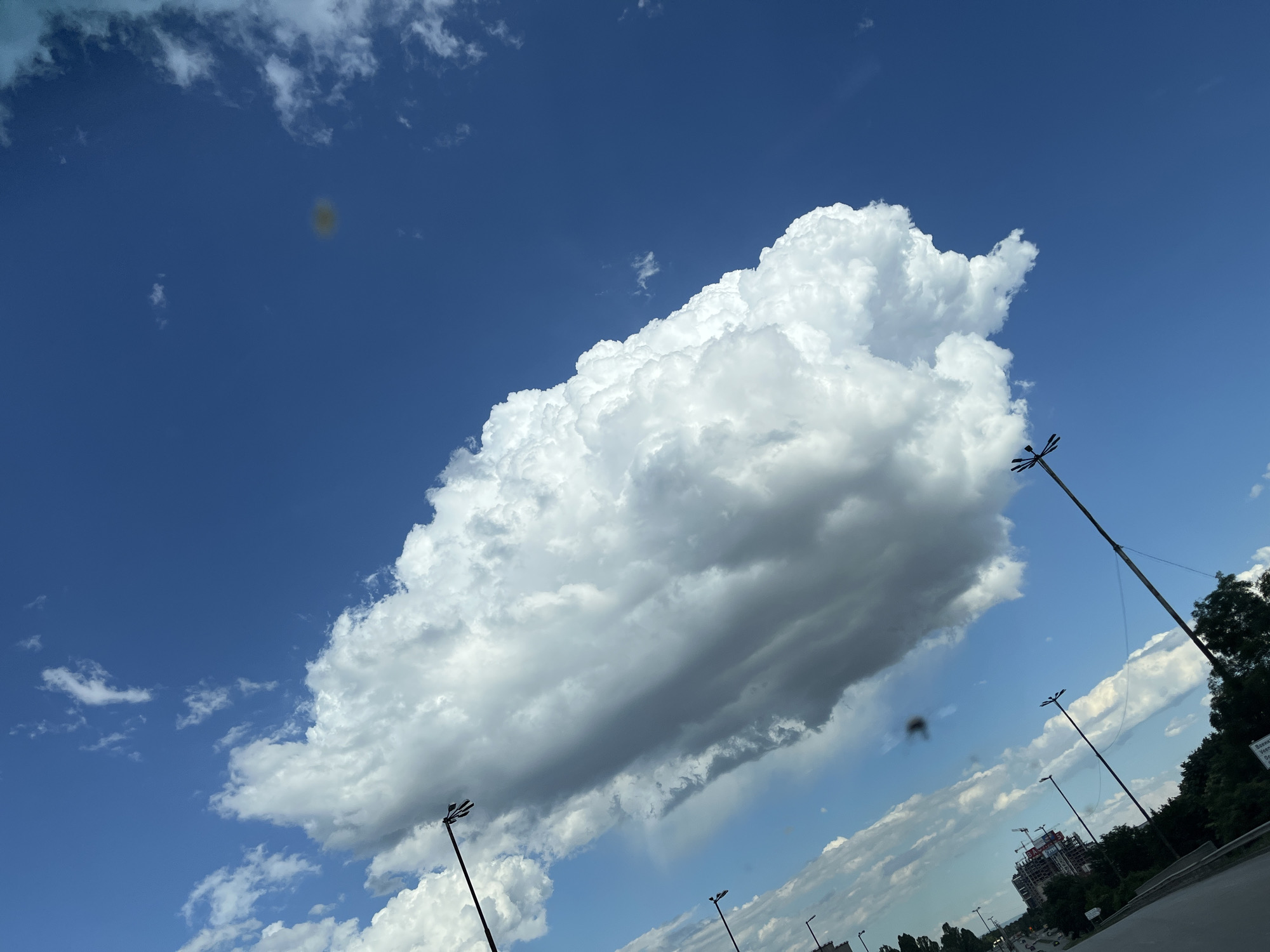 A wide-angle, low-angle shot captures a vast expanse of blue sky dominated by a large, fluffy white cumulus cloud. The cloud is positioned in the upper center of the frame, casting a slight shadow on the sky beneath it. In the foreground, a diagonal line runs from the bottom right to the upper left, suggesting a road or elevated pathway. Several black lampposts with multiple lights are scattered along this line, some tilted. To the far right, a dense line of dark green trees is visible, and beyond that, a multi-story building under construction with cranes on top. The sky also shows smaller, wispy white clouds scattered across it, and two dark circular smudges, possibly lens artifacts. The overall scene is bright and airy, with a focus on the sky and atmospheric conditions.