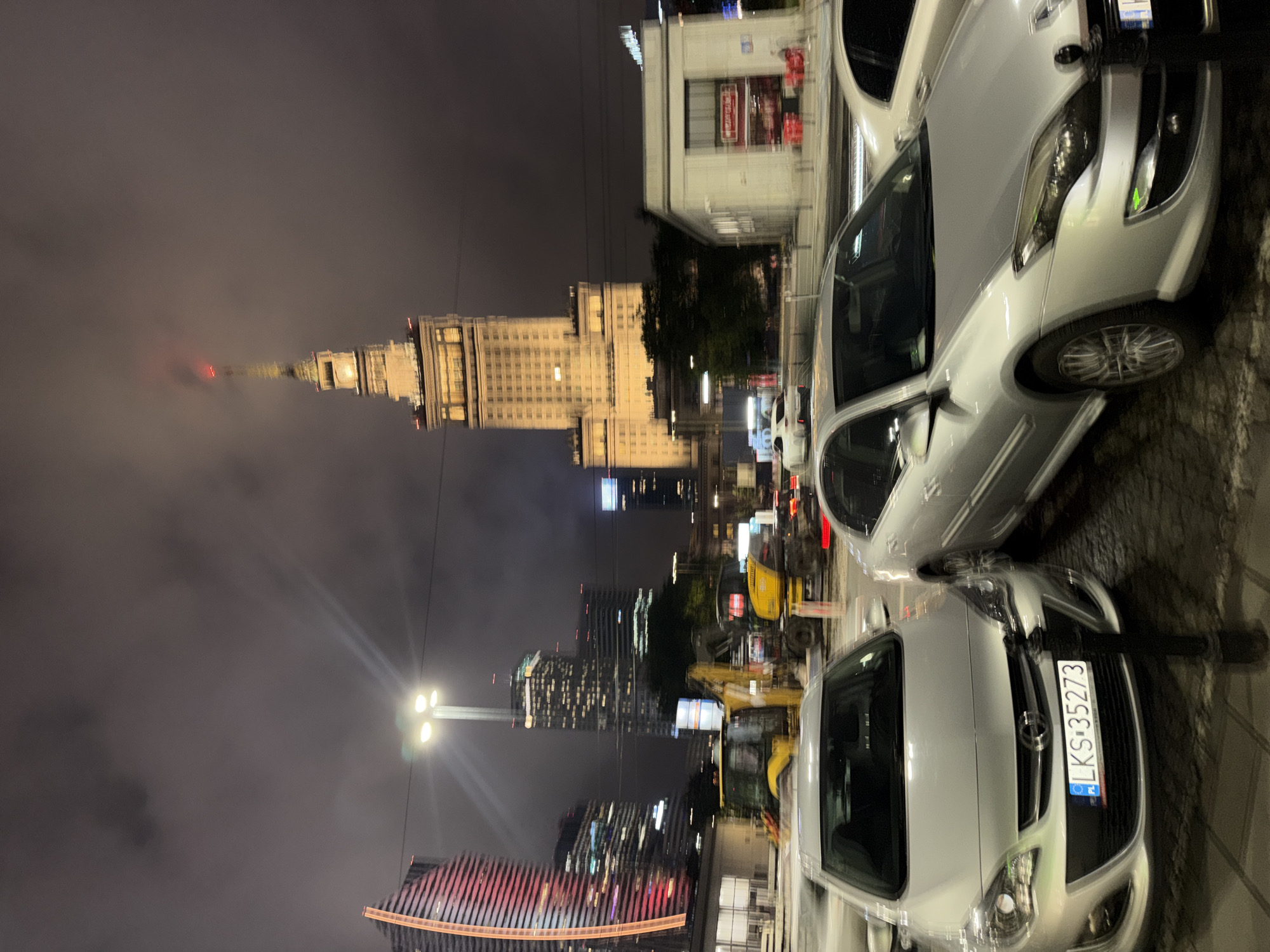 The image captures a street scene at night in a city, possibly Warsaw, Poland given the presence of a building resembling the Palace of Culture and Science. In the foreground, two silver cars are parked. The car on the left is a hatchback with a license plate reading "LKS 35273". The car on the right is also a silver sedan. Behind the cars, construction equipment, including yellow excavators, is visible. In the mid-ground, several modern high-rise buildings are illuminated. The most prominent structure is a tall, ornate tower in the center, lit up in yellow, with a red light at its peak, further suggesting it could be the Palace of Culture and Science. The sky is overcast and dark. The overall impression is one of a bustling urban environment at night, with construction and modern architecture dominating the landscape.
