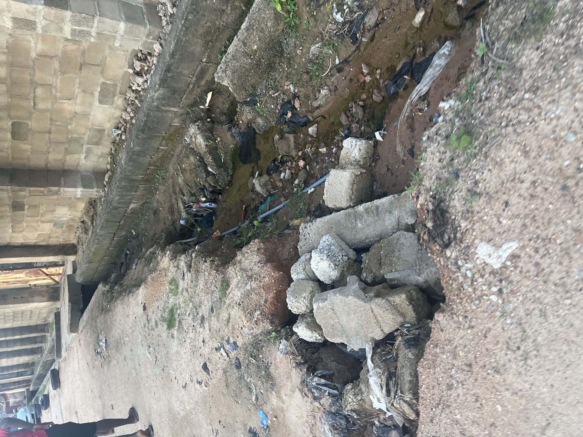 The image shows an outdoor scene with a focus on a drainage ditch filled with debris and some water. The ditch is lined with concrete and dirt, and there are many pieces of trash, including plastic bags and other refuse, scattered throughout. Some concrete blocks and rubble are piled up near the ditch. In the background, there appears to be a structure made of concrete blocks and possibly some scaffolding or beams, suggesting an urban or semi-urban environment.
On the left side of the image, a concrete walkway or path is visible, and there are a few people walking. One person in the foreground is wearing a red shirt and black shorts, with their back to the camera. Another person is partially visible further down the path. The ground is a mix of concrete and dirt. The overall impression is one of neglect and an accumulation of waste in a functional, but unmaintained, area.
The lighting suggests it is daytime, but the specific time of day is not clear. The weather appears to be overcast or at least not sunny, given the lack of strong shadows. There are no visible text elements in the image. The scene is consistent with the provided location context of Jalingo, Nigeria, depicting an area where waste management might be a challenge.
