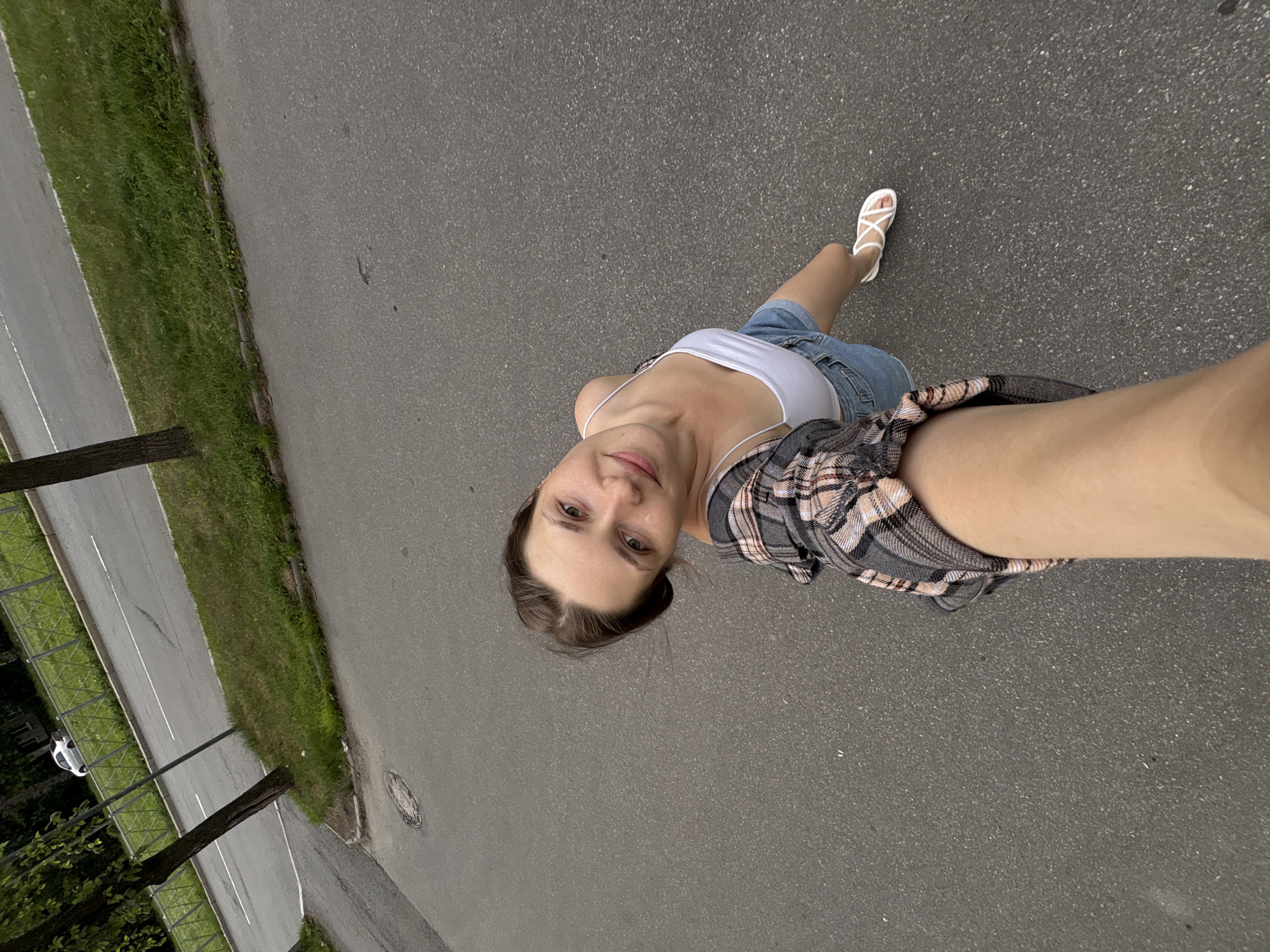 The image shows a young woman standing on an asphalt surface, possibly a road or pathway, in an outdoor setting. She is wearing a white tank top, denim shorts, and white sandals. A plaid shirt is draped over her shoulders. She is looking up towards the camera, her expression neutral. The scene includes a paved road with a white line marking, bordered by a grassy area and trees. There are also some distant streetlights visible. The lighting suggests it is daytime, with overcast weather. The presence of a manhole cover on the asphalt and the general appearance of the road and surrounding vegetation are consistent with an urban or suburban environment. No text is visible in the image.