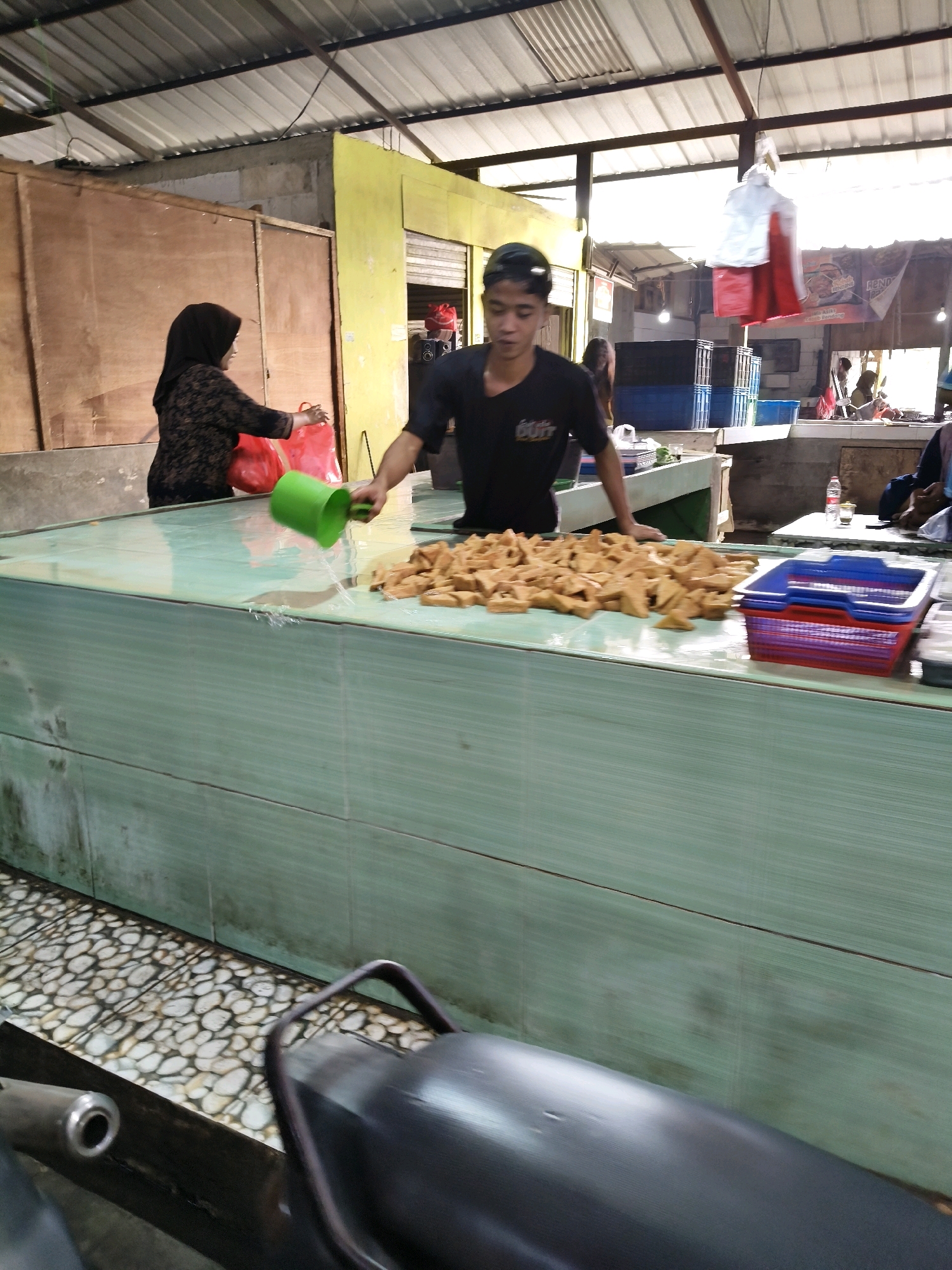 In Setiamekar, Indonesia, a young man in a black t-shirt and black baseball cap is pouring water from a green plastic pitcher onto a large display of fried food items, possibly tofu or tempeh, arranged on a light green tiled counter. The counter is wet, indicating the action is in progress. Behind him, a woman in a black hijab and patterned black top is holding two red plastic bags. The scene takes place in an open-air market with a corrugated metal roof supported by metal beams. In the background, there are other market stalls, including one with stacked blue crates and a banner. The floor is covered with a mosaic of light-colored pebbles. A motorcycle seat is visible in the foreground.