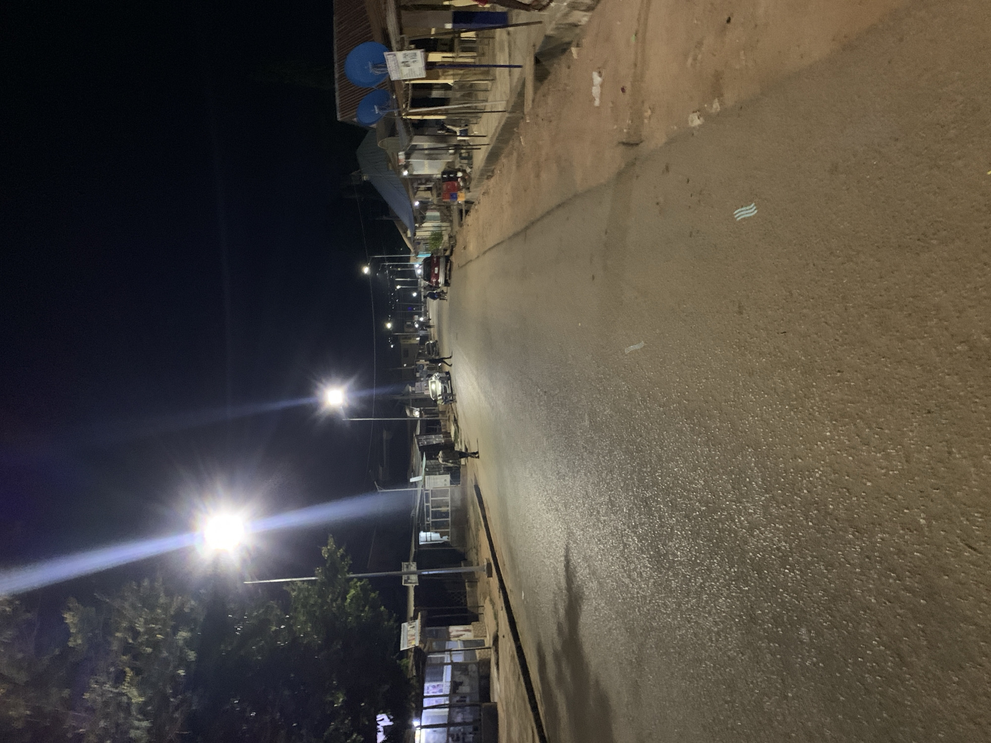 The image depicts a street scene at night in Jos, Nigeria. Streetlights illuminate a paved road lined with buildings on one side. The opposite side of the road is dark. Several people are walking along the street. A few parked vehicles are visible in the distance. The buildings appear to be small shops or stalls. The sky is dark. The overall impression is that of a quiet street at night.