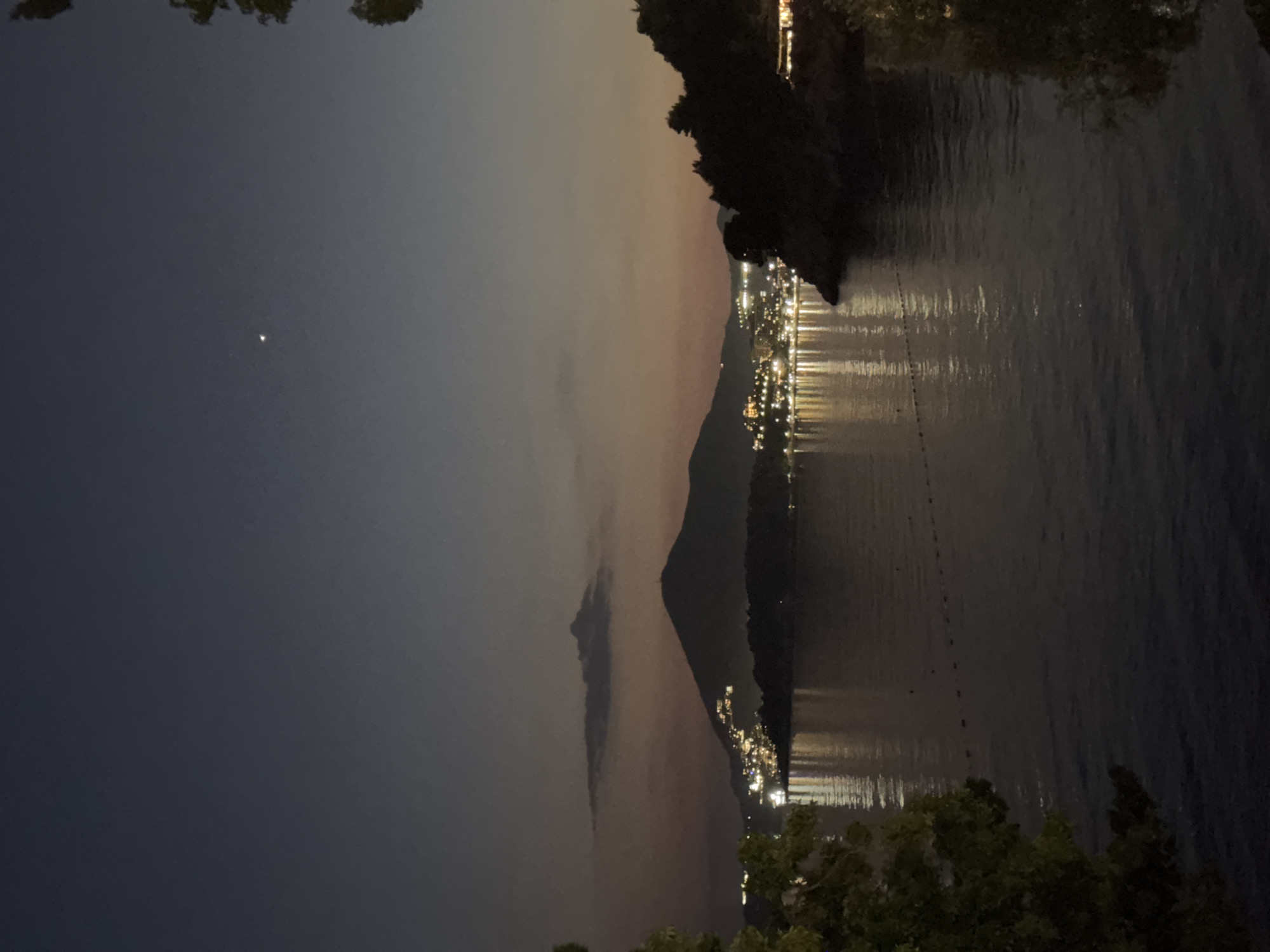 The image shows a nighttime view of a body of water, possibly a lake or bay, with a mountain silhouette in the background. There are lights visible on the shoreline, indicating a town or city. The sky is dark, with a faint light source, possibly the moon. Some trees are also visible in the foreground. The reflections of the city lights are visible in the water. The image is rotated 90 degrees clockwise. The exact location in Kamenovo, Montenegro, cannot be determined from the image, but the presence of a mountain and a body of water is consistent with the geography of the area.