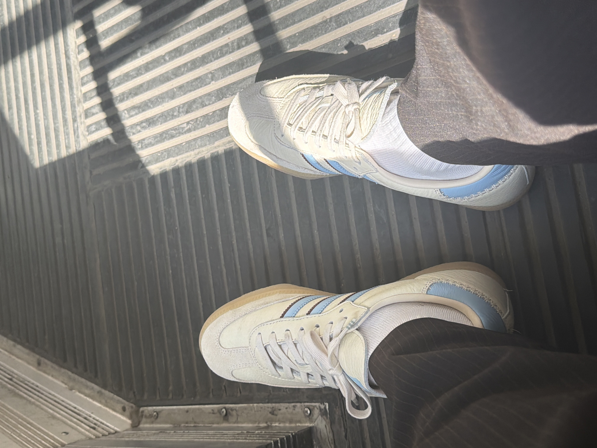 The image shows a downward-facing perspective of two feet wearing white Adidas Samba sneakers with light blue stripes and gum soles, standing on a grey, grooved metal floor. The legs are clad in pinstripe trousers. Light and shadow patterns cast stripes across the floor. The scene appears to be indoors. There's no clear indication that the location is Milan, Italy, or any other city. The image focuses solely on the footwear and immediate surroundings.