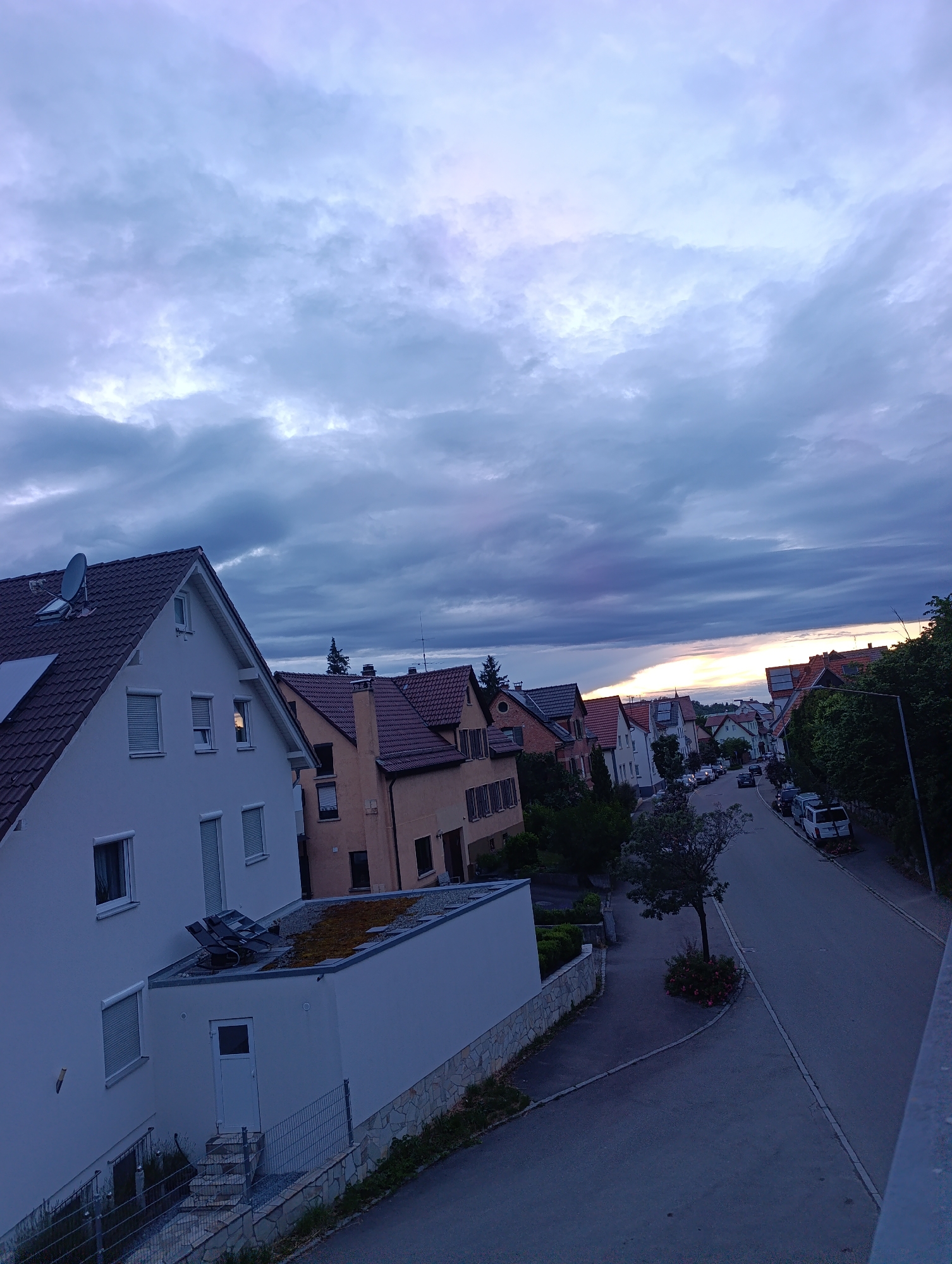 The image depicts a residential street in Pfullingen, Germany, at dusk. The sky is filled with dramatic clouds, tinged with purple and orange from the setting sun. On the left, a modern white house with a dark tiled roof dominates the foreground, featuring several windows and a satellite dish on the roof. Adjacent to it is another house with a similar roof, painted in a warm, muted orange hue.
The street curves gently to the right, lined with more houses, some with red-tiled roofs, and lush green trees. Several cars are parked along the street, suggesting a typical evening scene in a quiet neighborhood. A solitary tree stands in a small patch of greenery, surrounded by flowering bushes. The overall impression is one of a tranquil European residential area settling in for the evening.