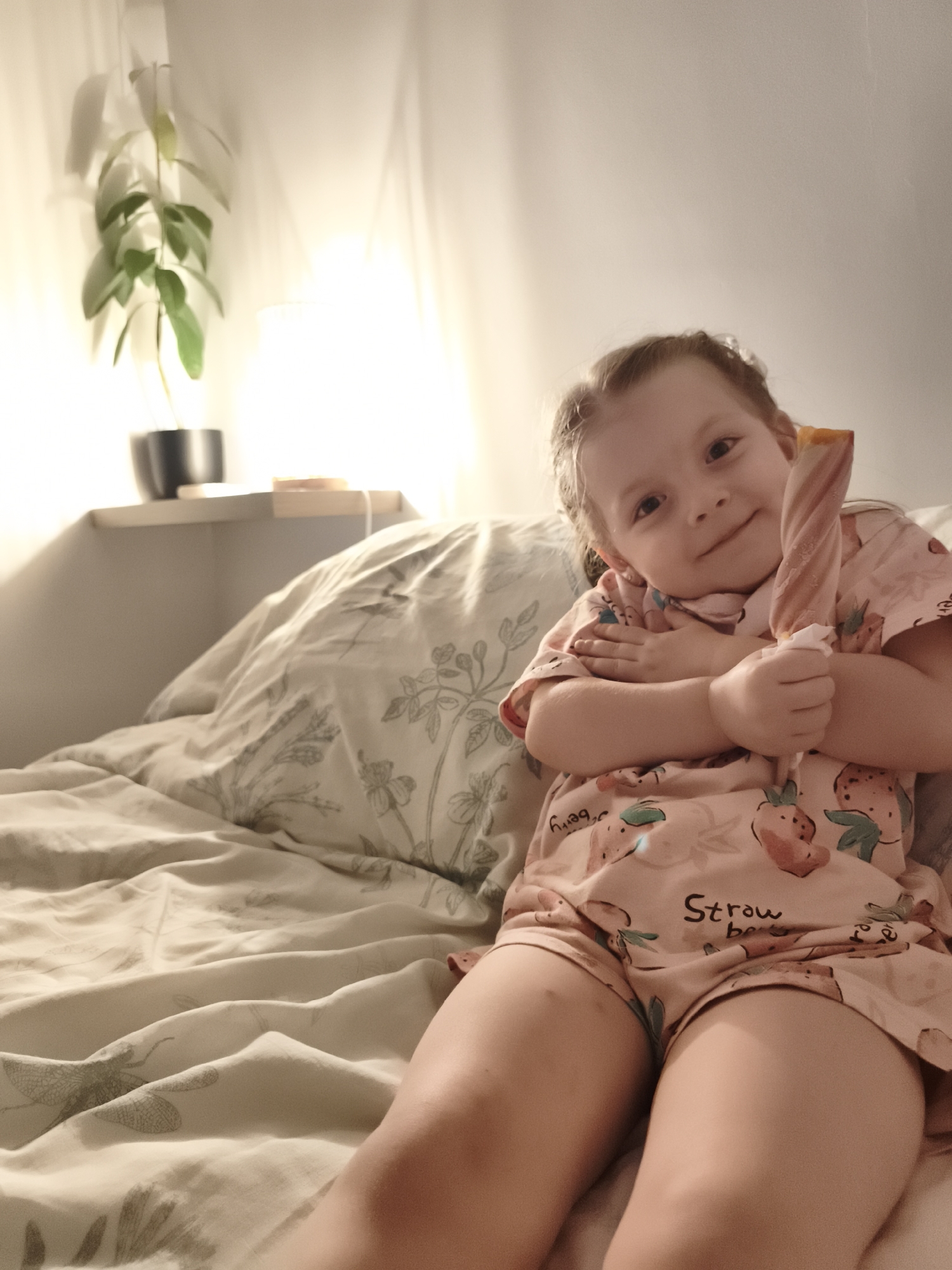 A young girl is sitting on a bed, holding a colorful popsicle. She is wearing a pink pajama set adorned with strawberries and the text "Strawberry". The bed is covered with a light-colored duvet featuring a floral and dragonfly pattern. In the background, a shelf holds a potted plant and a small wooden object. A soft light emanates from a lamp, casting shadows on the wall, suggesting it might be evening or a dimly lit room. The girl has a gentle smile and appears content. The setting seems to be a bedroom, possibly in Lagny-sur-Marne, France.