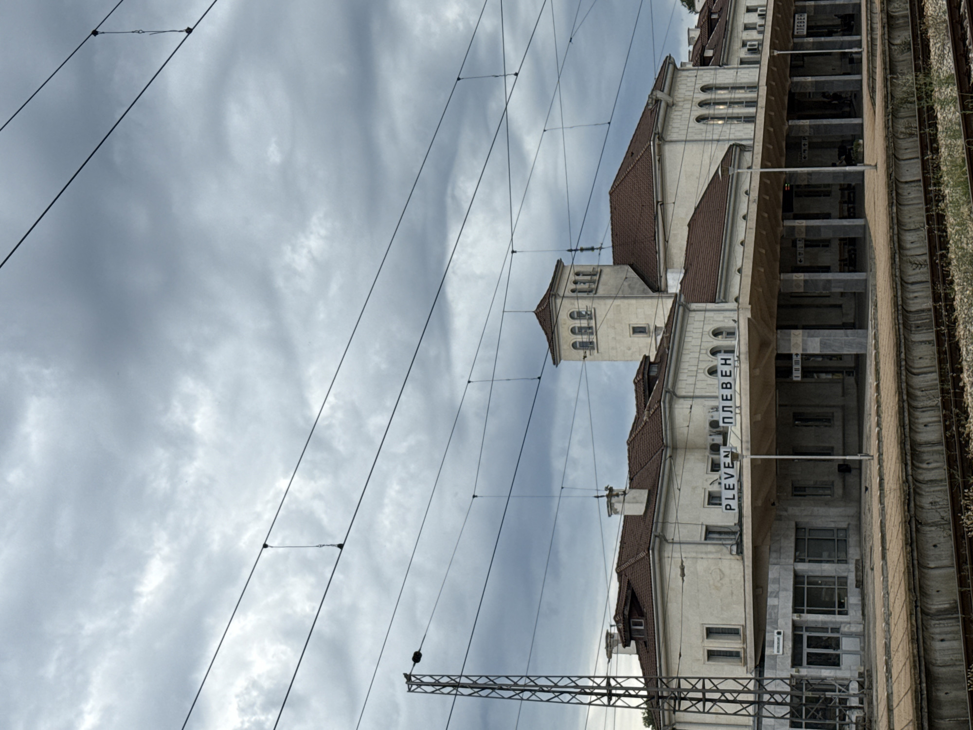 The image shows the Pleven railway station in Pleven, Bulgaria. The station building is a long, low structure with a covered platform in front. The building is made of light-colored stone and has a red tile roof. There is a clock tower on the right side of the building. The sky is cloudy and overcast. There are several train tracks in the foreground. Overhead, there are electrical wires for the trains. The name of the station, "PLEVEN", is visible on the building in both Latin and Cyrillic script.