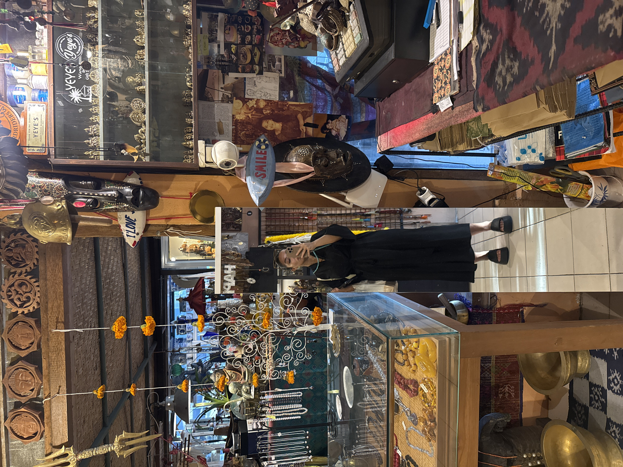 The image shows the interior of a shop in Kuta Selatan, Indonesia. A woman stands near a glass display case filled with jewelry, taking a selfie. Decorations, including orange flowers and other ornamental items, hang from the ceiling. Other items, such as wooden carvings, are displayed on the walls and on shelves. There are also signs with the shop's name, "Yeye's Waru Gift". Other merchandise such as clothing, trinkets, and decor are also displayed on shelves and tables within the shop.