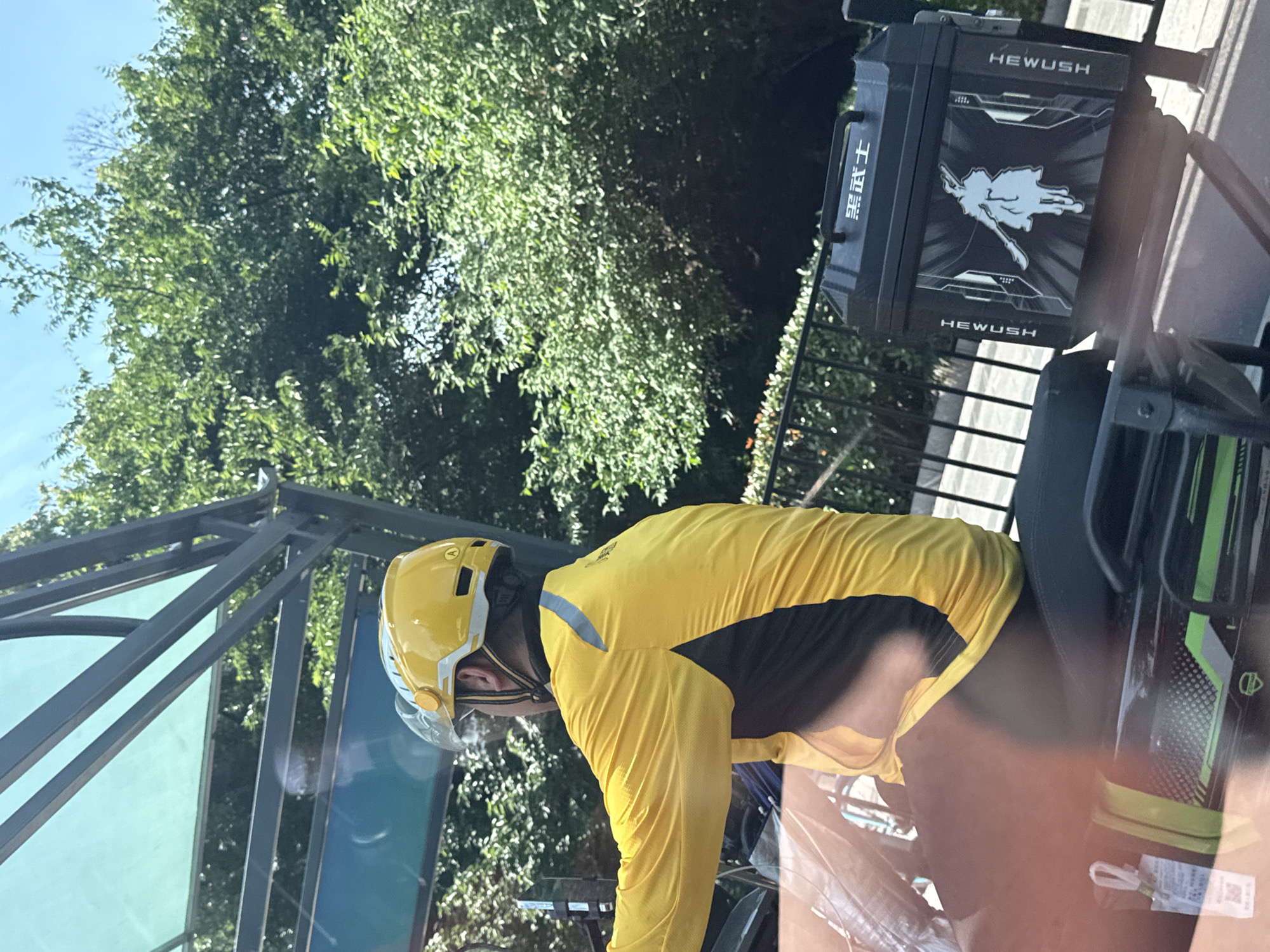 A person wearing a yellow and black long-sleeved shirt and a yellow helmet is riding a bicycle outdoors. The bicycle has a black box on the back with the word "HEWUSH" printed on it, along with Chinese characters "黑武士" and a white drawing resembling a figure with a sword. The surrounding environment includes lush green trees and a metallic structure overhead. The location is in the city of Gongshu District, China.