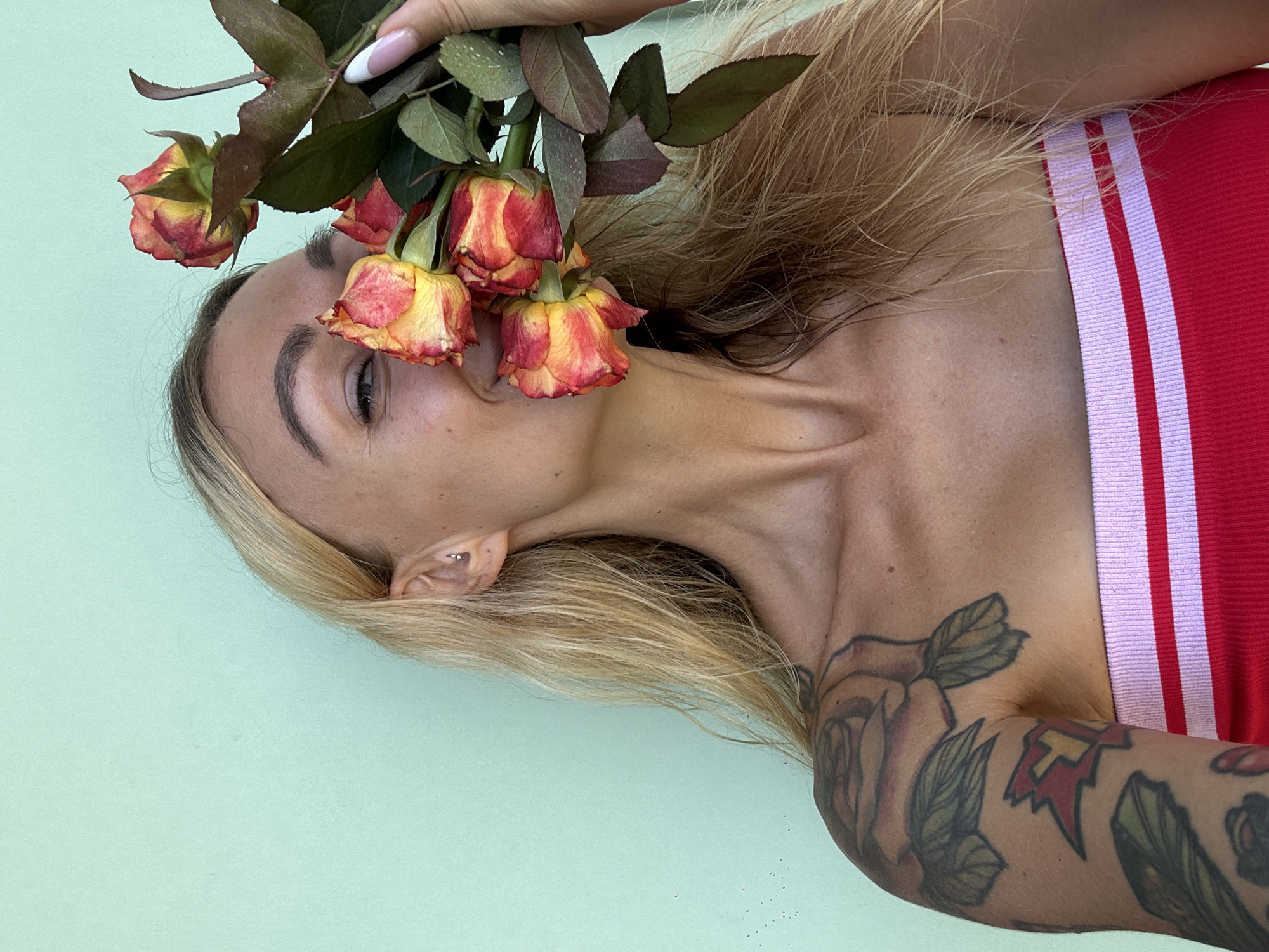 A blonde woman is holding a bouquet of roses in front of her face, obscuring her nose and mouth. She is wearing a red bandeau top with pink and white stripes. She has tattoos on her left arm and shoulder, including a large rose design. The background is a solid mint green wall. The lighting suggests it is daytime. The woman is likely in an outdoor setting, possibly a resort or a street in Mũi Né, Vietnam, given the tropical flora implied by the roses and the warm climate suggested by her attire.