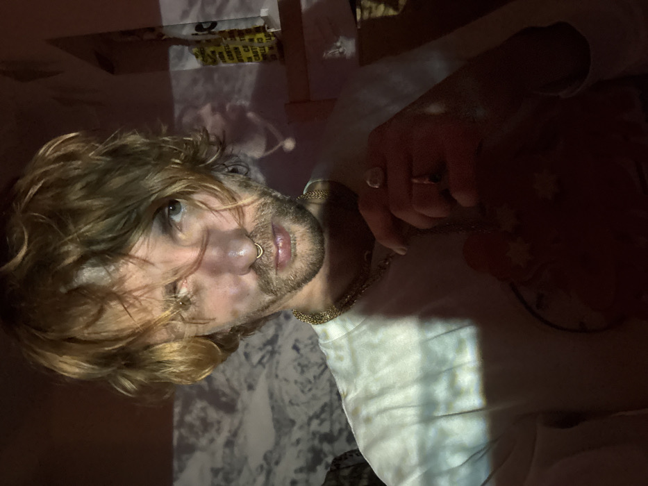 A person with shoulder-length, blonde hair, a nose ring, and a light beard gazes upwards. They wear a white shirt and gold necklaces and rings. A marble-like pattern is projected onto the wall and their shirt. The lighting is dramatic, casting shadows and highlights across their face and clothing. The background is dark and slightly out of focus. The image appears to be taken at an unusual angle.