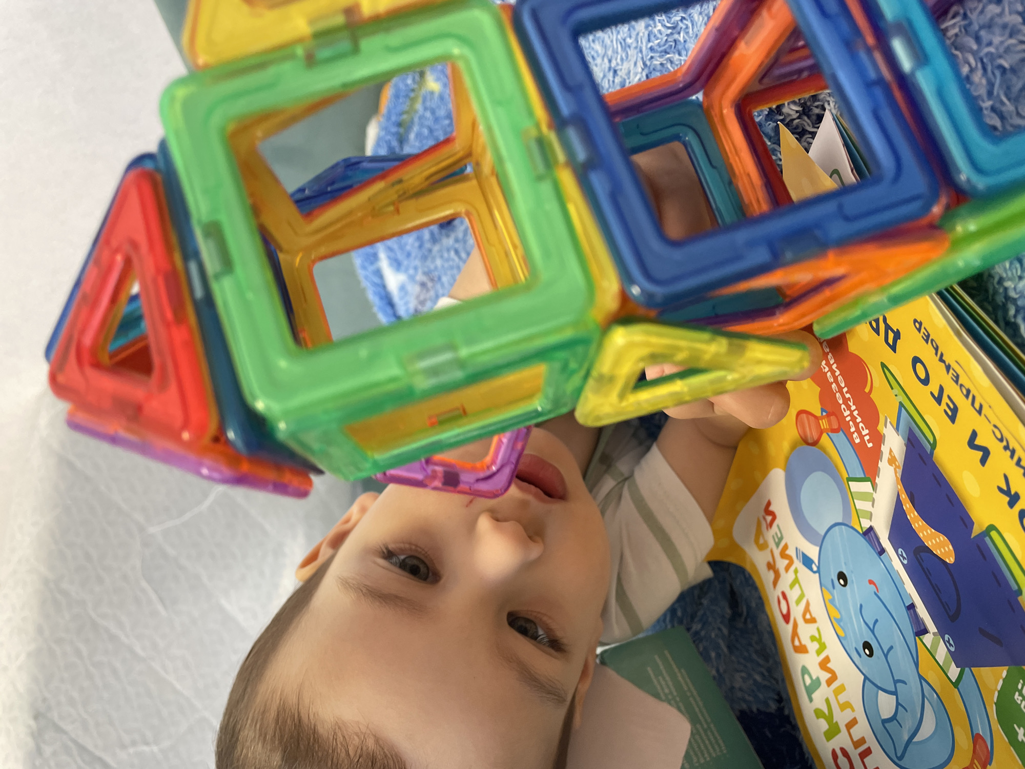 A young child is lying on a surface, possibly a bed or floor, amidst toys and books. The child is looking upwards, with colorful magnetic building blocks positioned above their face, forming a geometric structure. Several books surround the child, one of which is visible with a cartoon illustration of a blue elephant wearing a tie. The text on the book appears to be in Russian, indicating that the image could have been taken in a Russian-speaking region. There is no evidence in the image to confirm the location is Izhevsk, Russia.