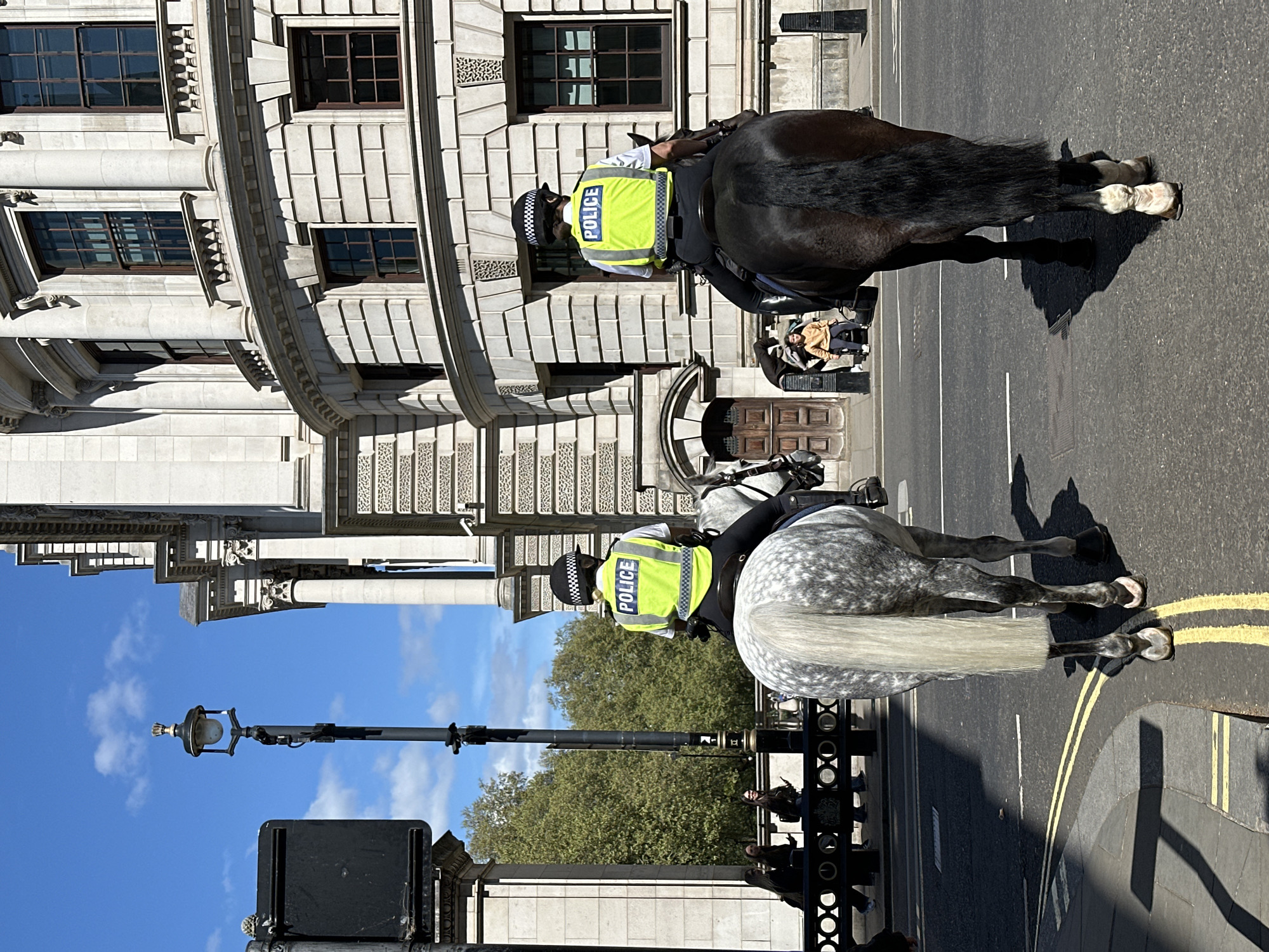 The image depicts two mounted police officers on patrol in London, United Kingdom. The officers are wearing high-visibility vests labeled "POLICE" and traditional helmets. They are riding a gray dappled horse and a dark horse, respectively, along a street marked with double yellow lines. A stone building with ornate architectural details is visible to the left. A lamppost and a wrought-iron fence line the street. Trees and a blue sky with scattered clouds are in the background. Several pedestrians are visible.