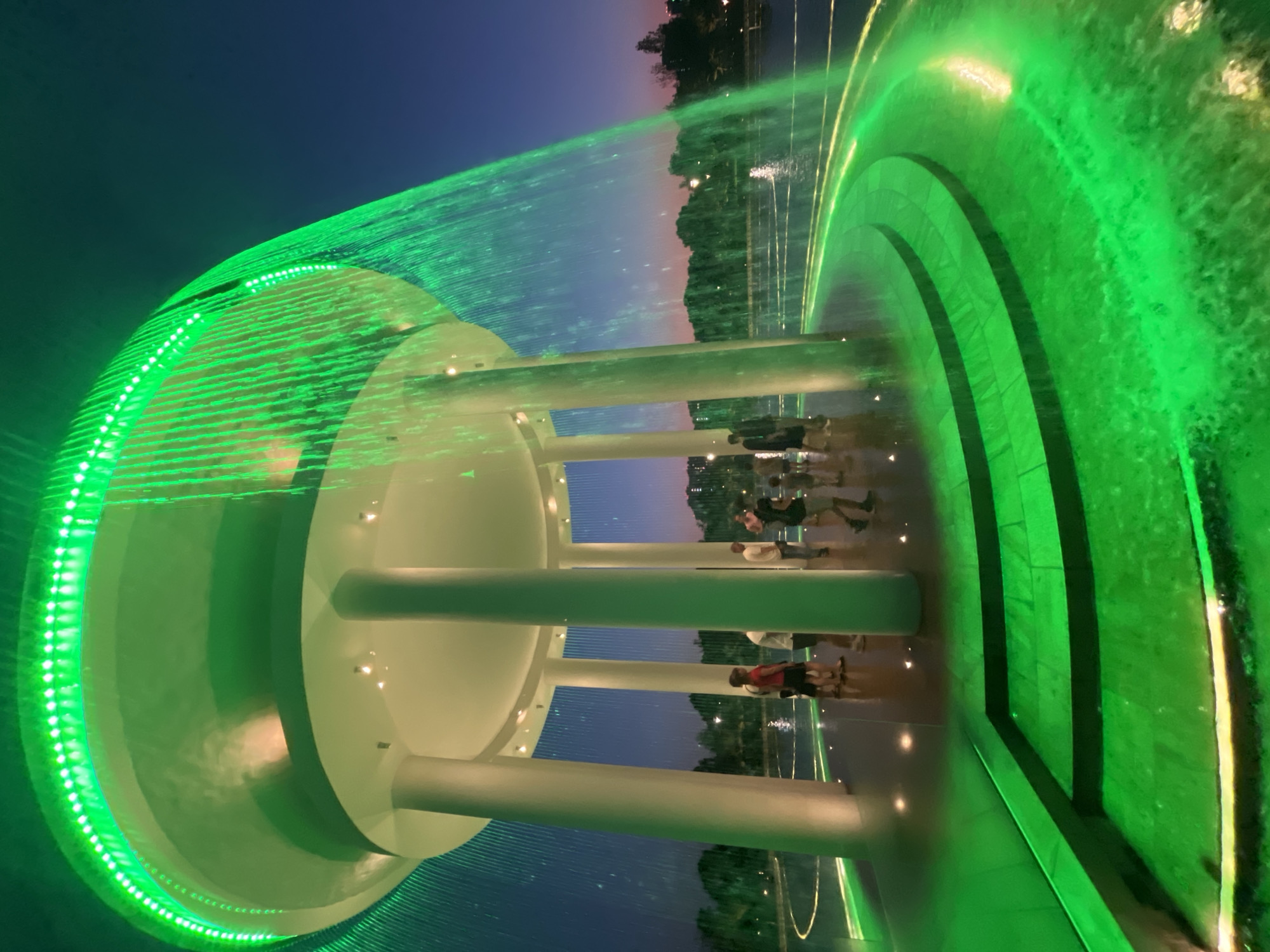 This media file captures a vibrant nighttime scene at a fountain, likely in Krasnodar, Russia, given the provided context. The fountain is illuminated with striking green lights, creating a dazzling spectacle. The water streams are caught in the light, appearing as iridescent curtains and arcs. A large, circular structure with white pillars forms the central part of the fountain's design, serving as a platform or viewing area.
Several people are visible, appearing as silhouettes or figures enjoying the display. They are casually dressed, suggesting a public space where people gather for leisure. Some are standing in groups, looking at the fountain, while others are walking. The ground around the fountain appears wet and reflective, enhancing the overall visual effect of the lights and water.
The time of day is clearly night, with a dark sky transitioning to hints of twilight colors near the horizon, where trees and distant structures are faintly visible. The overall mood is one of wonder and amusement, with the dynamic interplay of light and water creating an immersive and enchanting experience for the onlookers. No visible text is present in the media file.