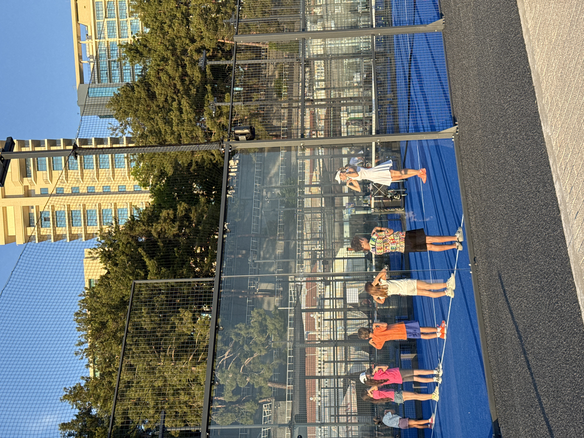A group of seven people, including children and adults, are standing in a line on a blue padel court. They appear to be participating in a training session or a game. The setting is outdoors, with a building and trees visible in the background, along with a chain-link fence surrounding the court. The time of day seems to be late afternoon, as indicated by the warm lighting and the long shadows. The people are dressed in athletic wear. One person is wearing a white dress, and others are in t-shirts and shorts. The mood appears to be focused and serious. There is no visible text in the image.