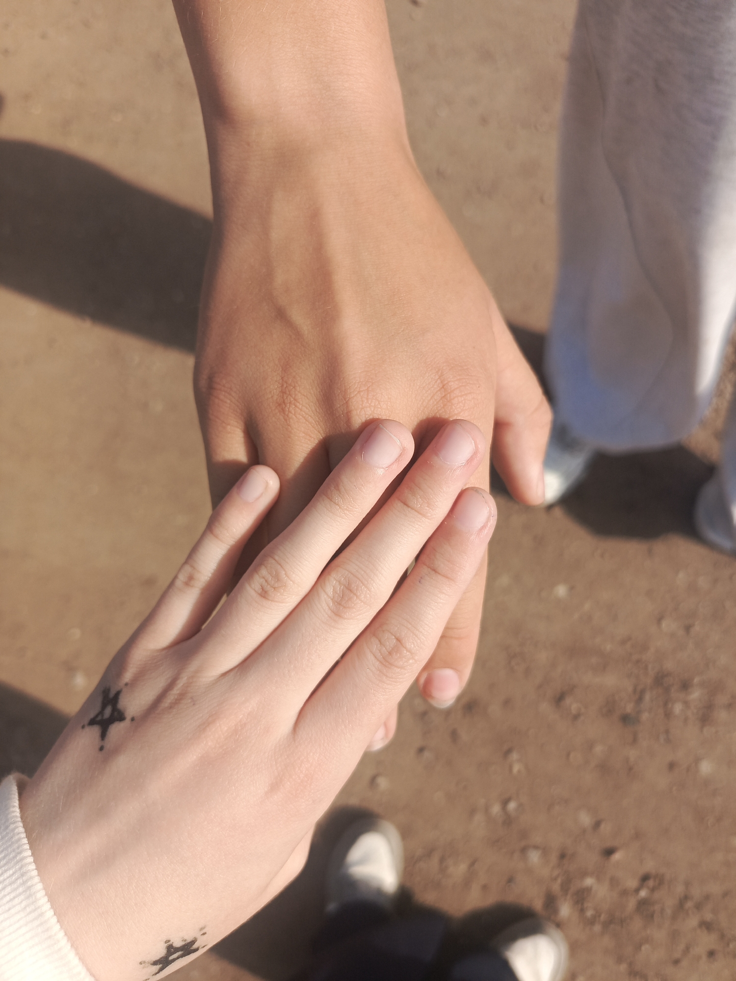 The image shows two individuals holding hands. The person on the left has lighter skin and a star tattoo on the back of their hand and a mark on their wrist. The person on the right has darker skin. Both individuals are wearing casual clothing. The ground is a tan, dirt-like surface. The location is Unknown, Unknown.