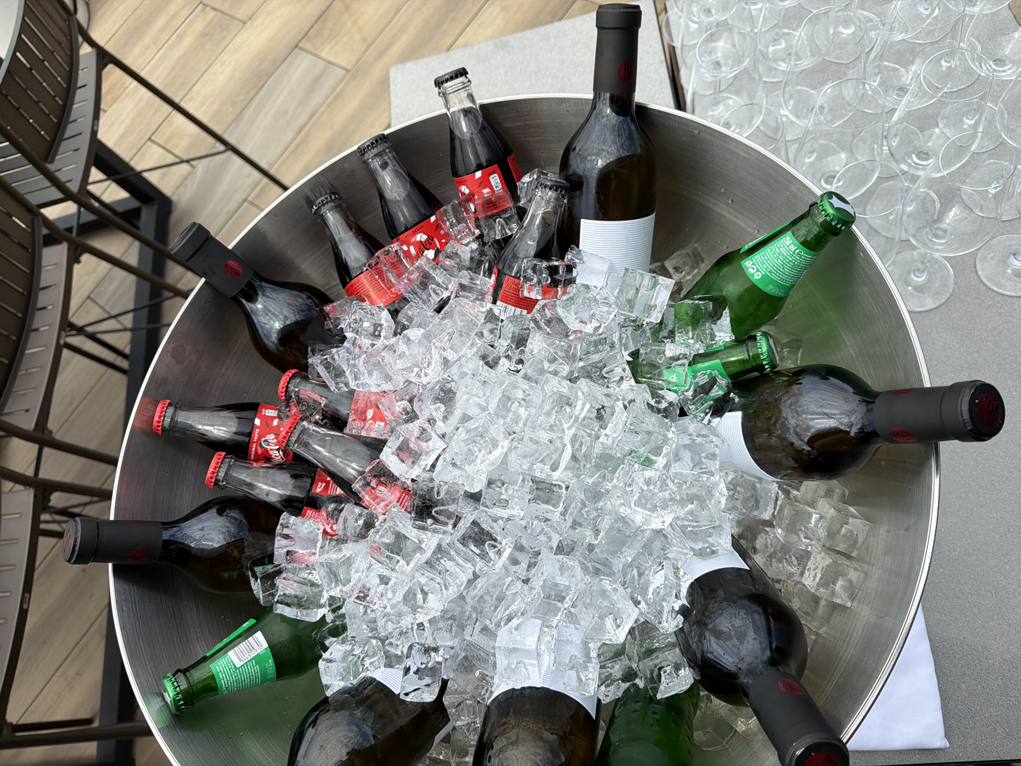 The image shows a round, metal ice bucket filled with ice and various beverages. These include several small glass bottles of Coca-Cola with red labeling, as well as wine bottles with dark glass and either white or red labels. Some wine bottles are green. There are many clear wine glasses near the ice bucket. The scene appears to be set up at an outdoor event, possibly a party or gathering. Metal chairs are visible to the left. It is not possible to tell the location in Sofia, Bulgaria from this image.