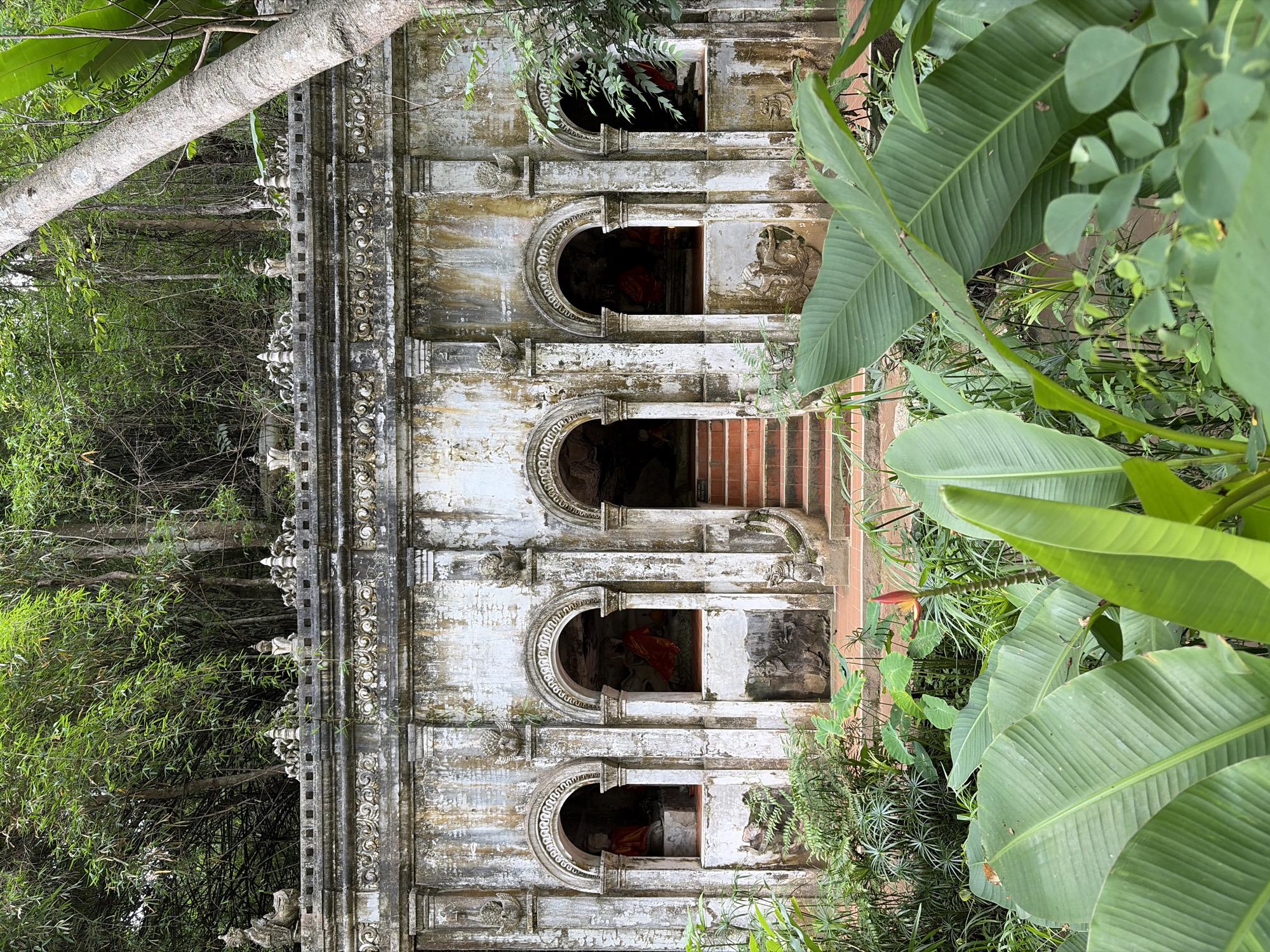 A stone facade of a building, possibly a temple or shrine, stands against a backdrop of lush green foliage and trees. The facade features multiple arched alcoves, each housing a statue of Buddha adorned with orange fabric. The stone structure is weathered and stained, indicating its age and exposure to the elements. Decorative carvings adorn the upper sections of the facade. In the foreground, large, vibrant green leaves of tropical plants dominate the lower portion of the image. A staircase with red treads leads up to one of the central arched entrances. The scene suggests a serene and somewhat overgrown spiritual site nestled within a natural environment. This site is located in the city of Suthep Town Municipality, Thailand.