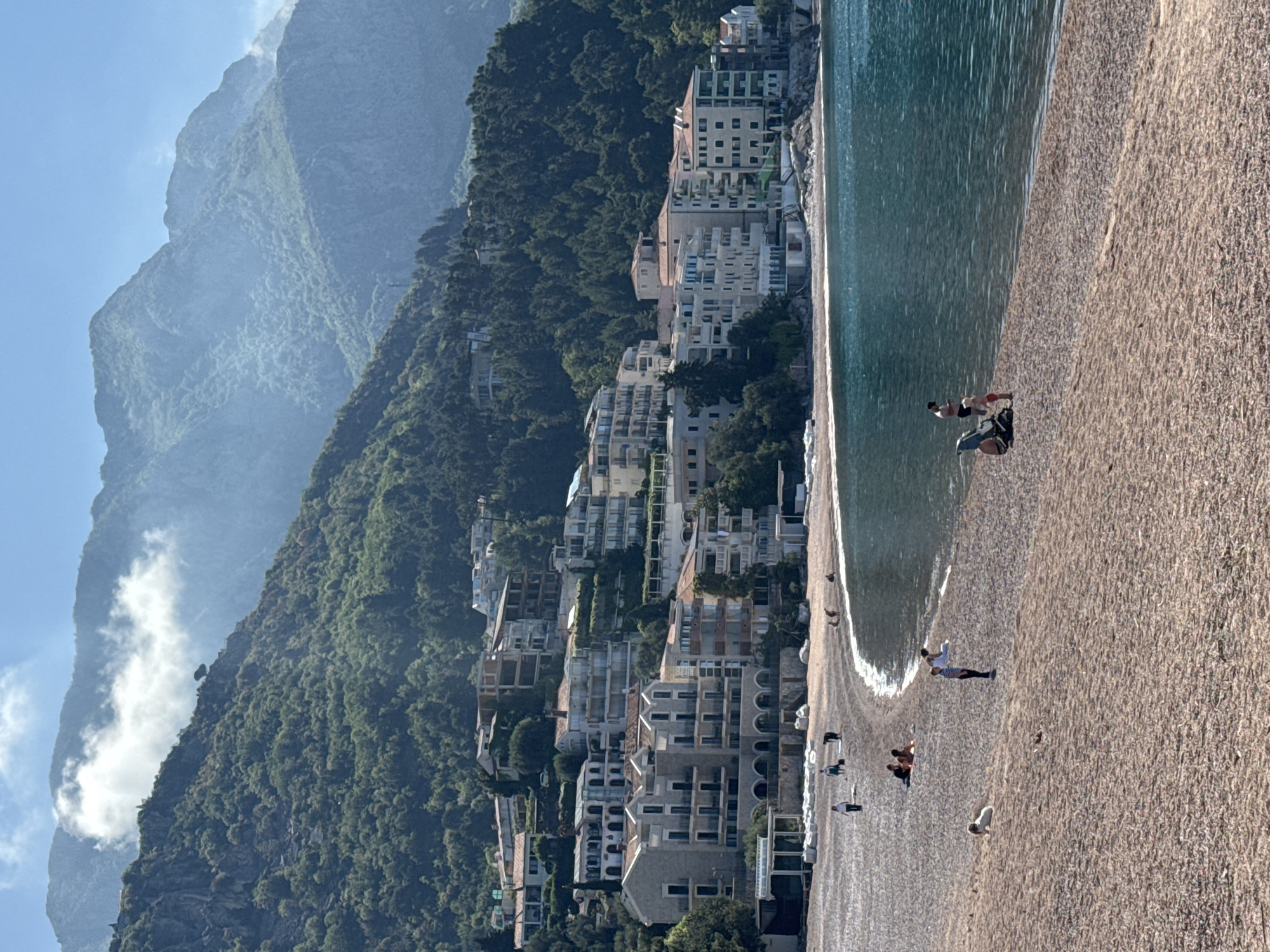 The image shows a beach in Sveti Stefan, Montenegro. The beach is lined with buildings along the coast. In the background are large mountains partially obscured by clouds. People are visible on the beach, some standing and others relaxing. The sea is calm with gentle waves lapping at the shore.