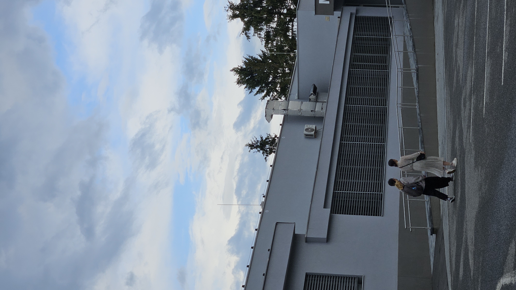 The image shows two young adults walking away from the camera across a paved area in front of a modern, gray building. The building has long horizontal windows or vents. To the right of the building, a portion of a white sign with a dark letter "M" is visible. In the background, several evergreen trees are visible against a cloudy sky. One person, wearing dark pants, a striped shirt, and a backpack, walks slightly ahead of the other person, who wears a light-colored dress and a dark crossbody bag. The pavement has white lines marking parking spaces. The scene appears to be outdoors in an urban or semi-urban environment. The overall lighting suggests it is daytime, possibly late afternoon or an overcast day.