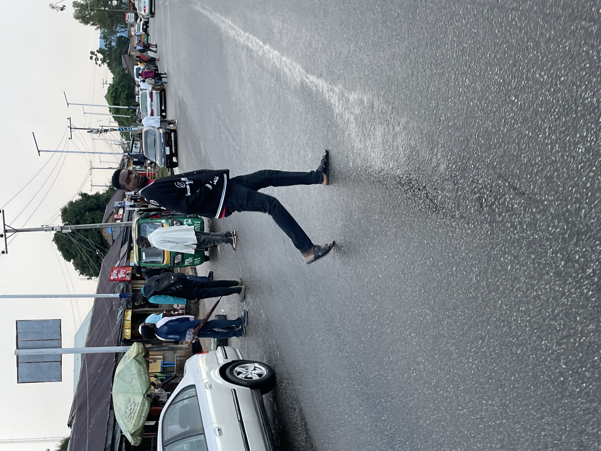 A man is walking across a wet, paved road in Jos, Nigeria. He is wearing a black jacket with white lettering, black pants, and dark sandals. He is looking towards the camera. Behind him, several other people are visible, some standing near a green and yellow auto-rickshaw. A silver car is parked on the left side of the road. In the background, there are buildings with corrugated metal roofs, streetlights, and trees. The sky is overcast. The scene appears to be a commercial or roadside area.