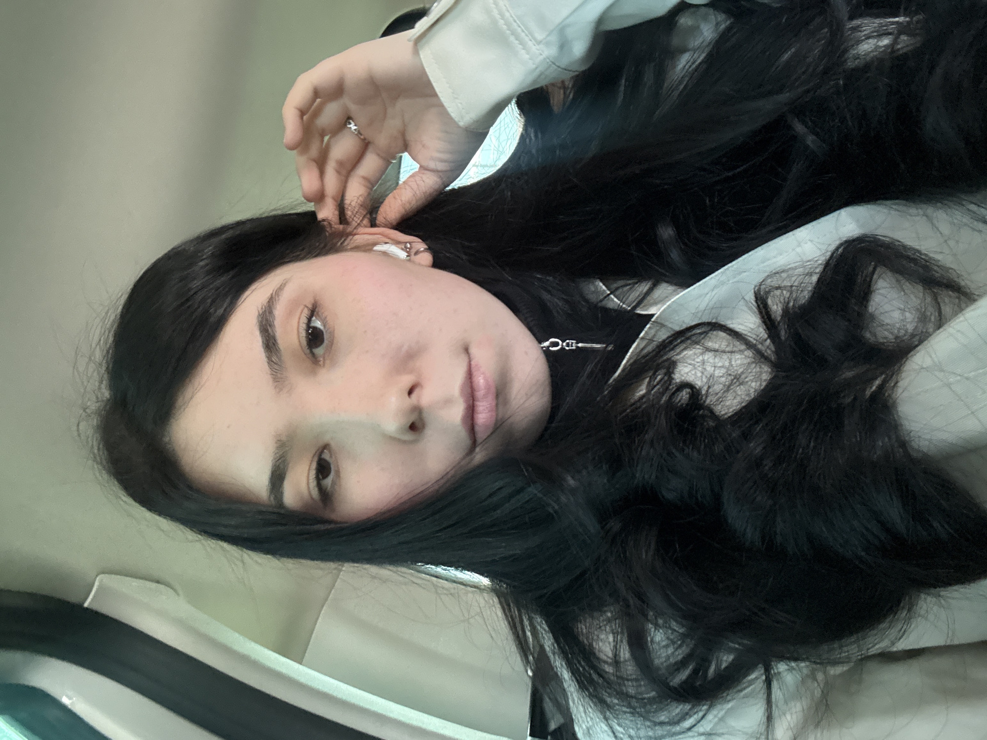 A person with long, dark hair is shown in a selfie-style photograph. The person is wearing a light-colored jacket and appears to be wearing a white wireless earbud in their left ear. Their left hand is raised near their ear. The background is indistinct but appears to be the interior of a vehicle. There is no discernible information indicating a specific location in Naberezhnye Chelny, Russia.