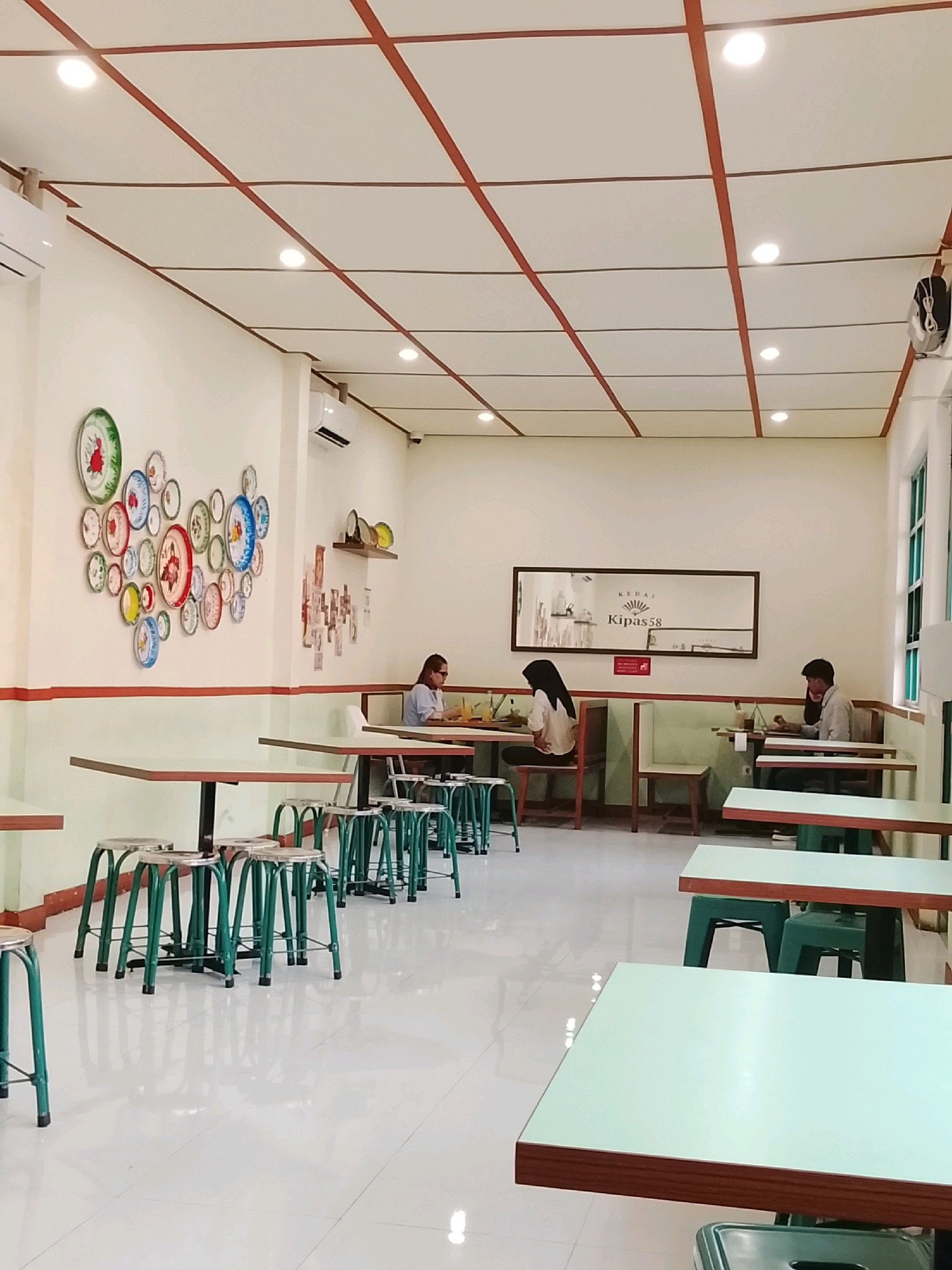 This is a full shot of the interior of a cafe called "Kipas58" located in Surabaya, Indonesia. The cafe has a modern, minimalist design with white walls and a white tiled floor. The ceiling is white with brown wooden beams forming a grid pattern, and round lights are evenly spaced throughout.
On the left wall, there's a decorative arrangement of colorful circular plates in various sizes, creating a whimsical focal point. Two people are seated at tables. One woman, wearing a light-colored top and glasses, is seated at a table in the middle left of the frame, facing away from the camera. She appears to be looking at something on the table. Further back and to the right, a man wearing a grey shirt is seated at another table, also facing away from the camera. He is leaning forward, possibly engaged with something on his table. Another person, a woman wearing a black hijab and a light-colored blouse, is seated at a table in the center, with her back to the camera, seemingly engaged in a task at her table.
The cafe is furnished with light-colored tabletops with wooden edges, supported by dark legs. In the foreground and extending into the middle ground, rows of green metal stools with round silver seats are neatly arranged around the tables. The reflective floor creates a bright and spacious atmosphere. A large rectangular mirror hangs on the back wall, reflecting the interior and displaying the cafe's name, "KIPAS58". A window with multiple panes is visible on the right side of the frame. The overall environment appears calm and orderly, with patrons engaged in individual activities.