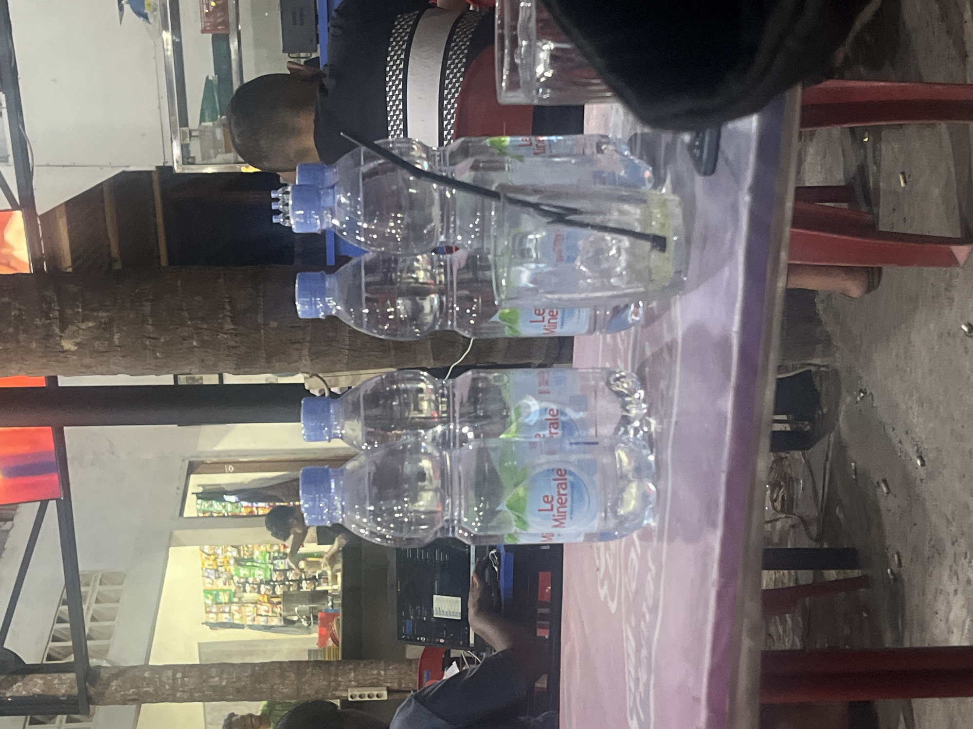 The image depicts an outdoor seating area, possibly at a street food stall or a casual eatery in Banda Aceh, Indonesia. In the foreground, a pink plastic table holds several partially filled bottles of "Le Minerale" brand water and an empty glass with a black straw. The table's surface is slightly reflective. Behind the table, on the ground, there are scattered cigarette butts.
In the middle ground, a person's back is visible, with their torso facing away from the camera and towards the left side of the frame. They appear to be wearing a black shirt with a white checkered pattern across the shoulders and chest. To the left of this person, a laptop is open on a table or counter, and someone's hands are visible, actively typing on the keyboard. Further left, in the background, a man in a green shirt is partially visible, smiling and looking towards the left.
The environment suggests a warm climate, with a large tree trunk visible on the right side of the frame, partially obscuring the view of a building in the background. This building has an open-fronted section displaying racks of goods, possibly snacks or drinks, reminiscent of a small convenience store or warung. The overall lighting is dim, indicating it is likely evening or night. The scene conveys a relaxed, informal atmosphere.
