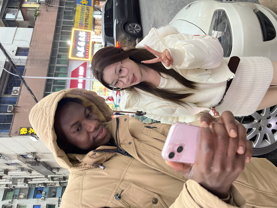 Two individuals, a male and a female, are depicted in an urban street setting in Jiangjin, China, posing for a selfie. The male, positioned on the left and appearing to be of African descent, wears a tan-colored hooded winter jacket and holds a light pink smartphone with a dual-camera system in his right hand, directed towards both subjects. He has a slight smile and dark, short hair. The female, positioned to the male's right and appearing to be of East Asian descent, wears a white puffer jacket over a white ribbed knit dress and round, light-rimmed eyeglasses. She displays a slight smile and makes a V-sign with her raised left hand. The background consists of multi-story buildings characteristic of an urban environment, with illuminated commercial signs displaying Chinese characters, including "仙居饭店" (Xianju Fandian) and "雅文." A white car is partially visible in the lower right foreground. The scene is illuminated by artificial light, suggesting nighttime or dusk, and the subjects' heavy clothing indicates cool weather conditions.