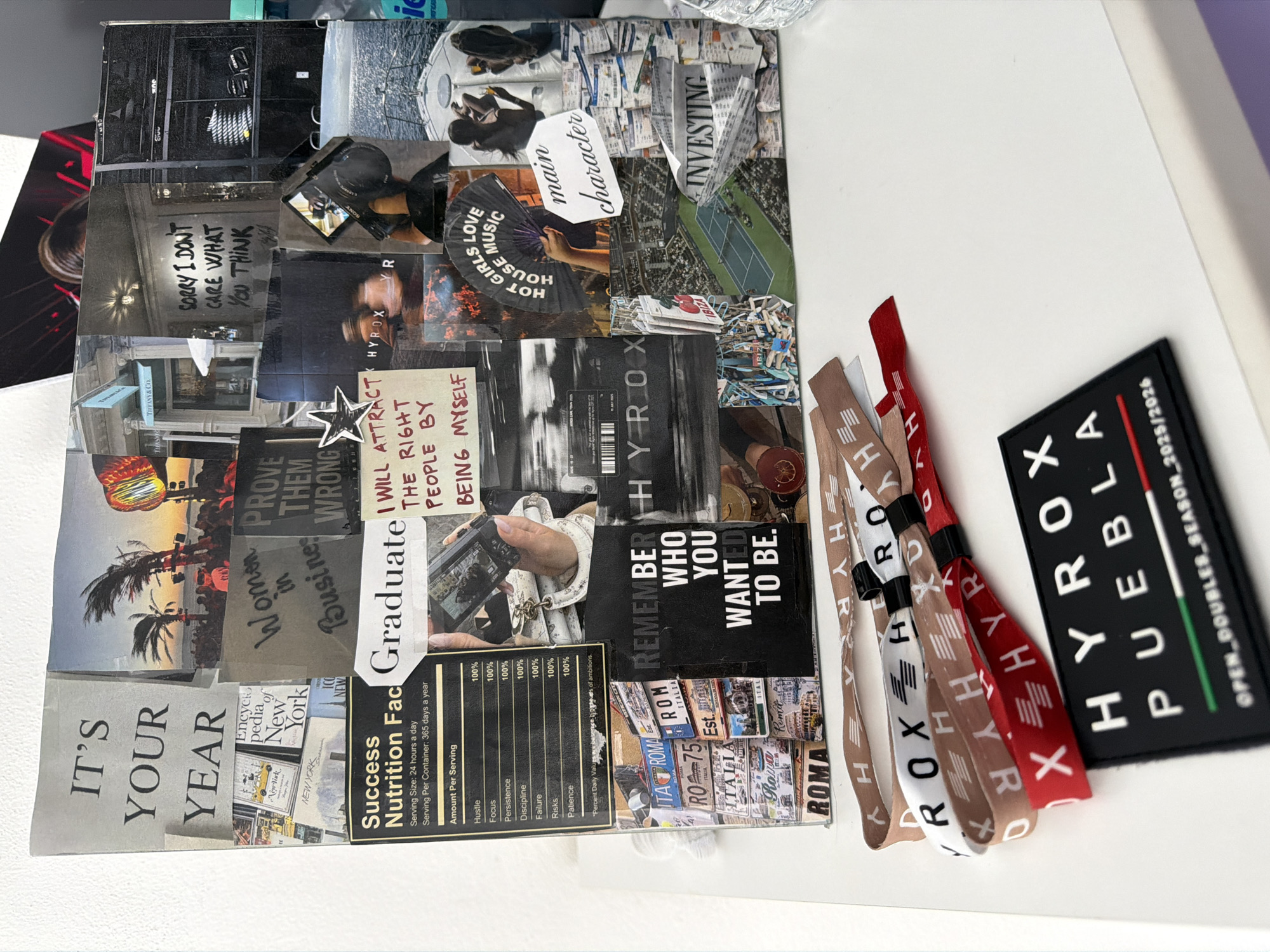 The image shows a collage board with motivational images and text. Key phrases include "It's Your Year," "Prove Them Wrong," "I Will Attract the Right People by Being Myself," and "Remember Who You Wanted to Be." Also present are images suggesting travel to New York and Rome, along with a "Success Nutrition Facts" label.
To the right of the collage are several HYROX wristbands in various colors (brown, white, and red), and a black HYROX PUEBLA patch with the text "OPEN_DOUBLES_SEASON_2025/2026" on it. Puebla is a city in Mexico. The scene is likely situated on a white table or surface. There are no people directly visible in the image, but there are implied subjects within the collage, such as businesswomen and graduate students. The overall environment appears to be indoors, with neutral lighting.