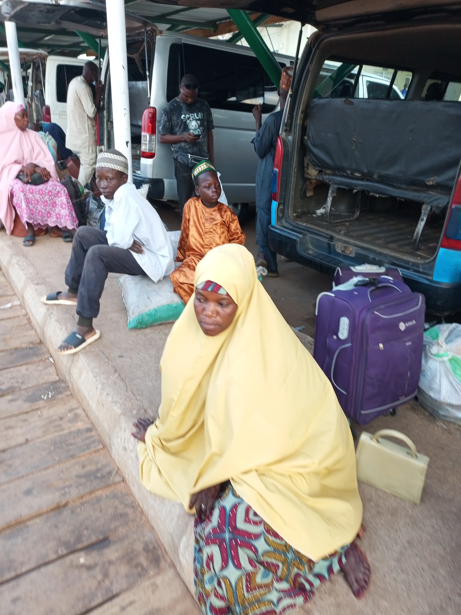In Gombe, Nigeria, a group of people are gathered near a parked vehicle. The scene appears to be an informal waiting area, possibly a bus stop or a transport hub. Several individuals are seated on the ground or on cushions, suggesting they are waiting for transportation.
In the foreground, a woman wearing a yellow hijab and a patterned skirt is seated, with her face partially visible. To her left, a young boy in an orange outfit and a green cap is also seated. To the left of the boy, a young man in a white shirt and dark pants is seated on a cushion. Behind them, a woman in a pink hijab and a patterned dress is seated. Several other people are visible in the background, some standing and some seated near vehicles.
Objects in the scene include luggage, with a prominent purple suitcase placed next to the woman in the yellow hijab. There are also bags and other personal belongings scattered around. The environment suggests an outdoor setting, with a concrete curb and a wooden surface visible in the foreground. A parked vehicle, likely a van or minibus, is the dominant object in the background, with its rear door open. The overall impression is one of people waiting, possibly for a journey, in a functional but not highly organized transportation area.