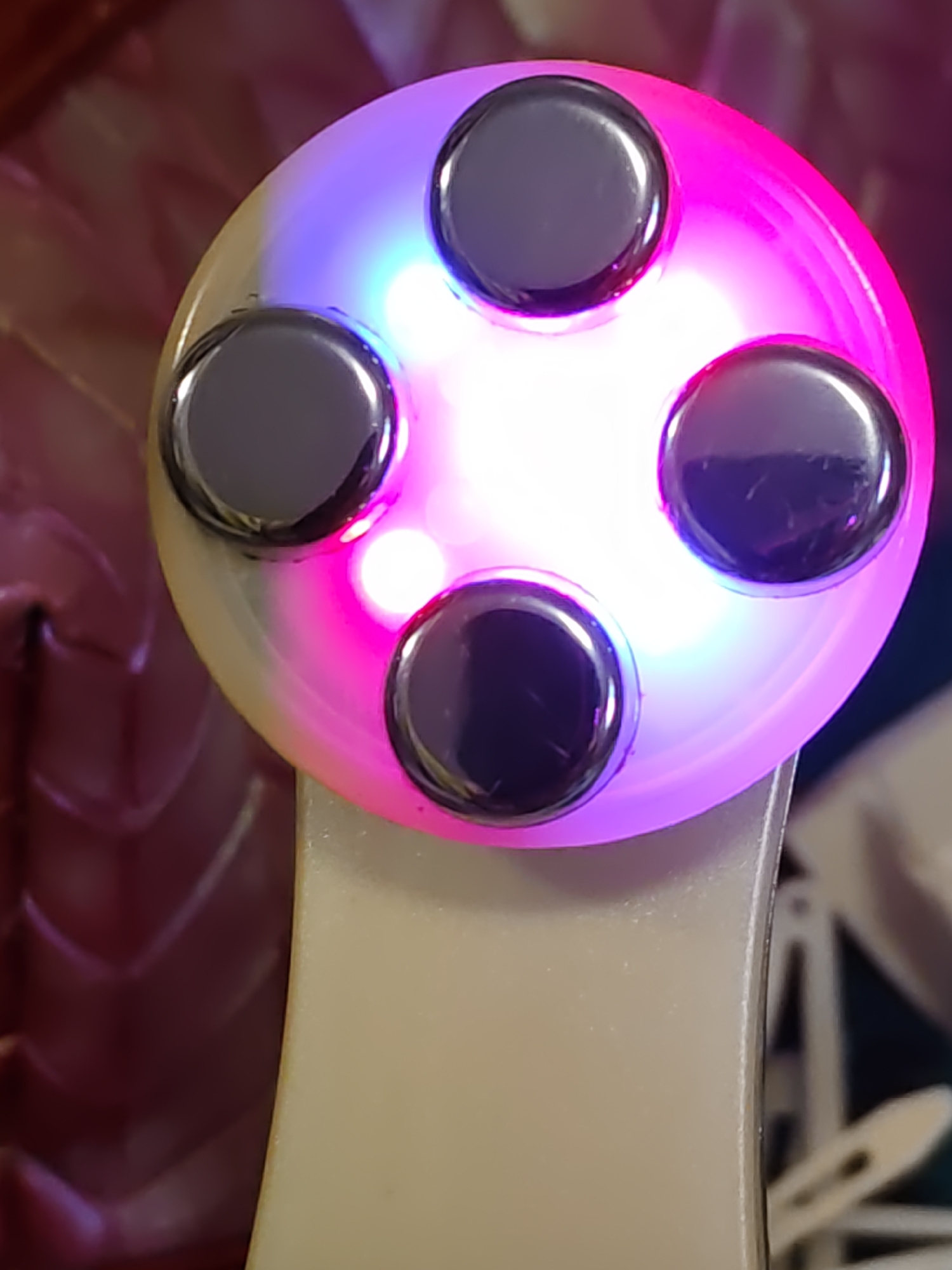 The image shows a close-up of a white beauty device with four metallic circular nodes arranged in a diamond pattern on a circular head. The head of the device is illuminated with pink and blue LED lights. The device is held at an angle, and its cream-colored handle is visible at the bottom. The background is blurred, with shades of pink and purple suggesting a soft, out-of-focus texture. The lighting suggests a studio or indoor setting, possibly a beauty salon or a home environment. There are no people or any context indicating its location in Rostov-on-Don, Russia.