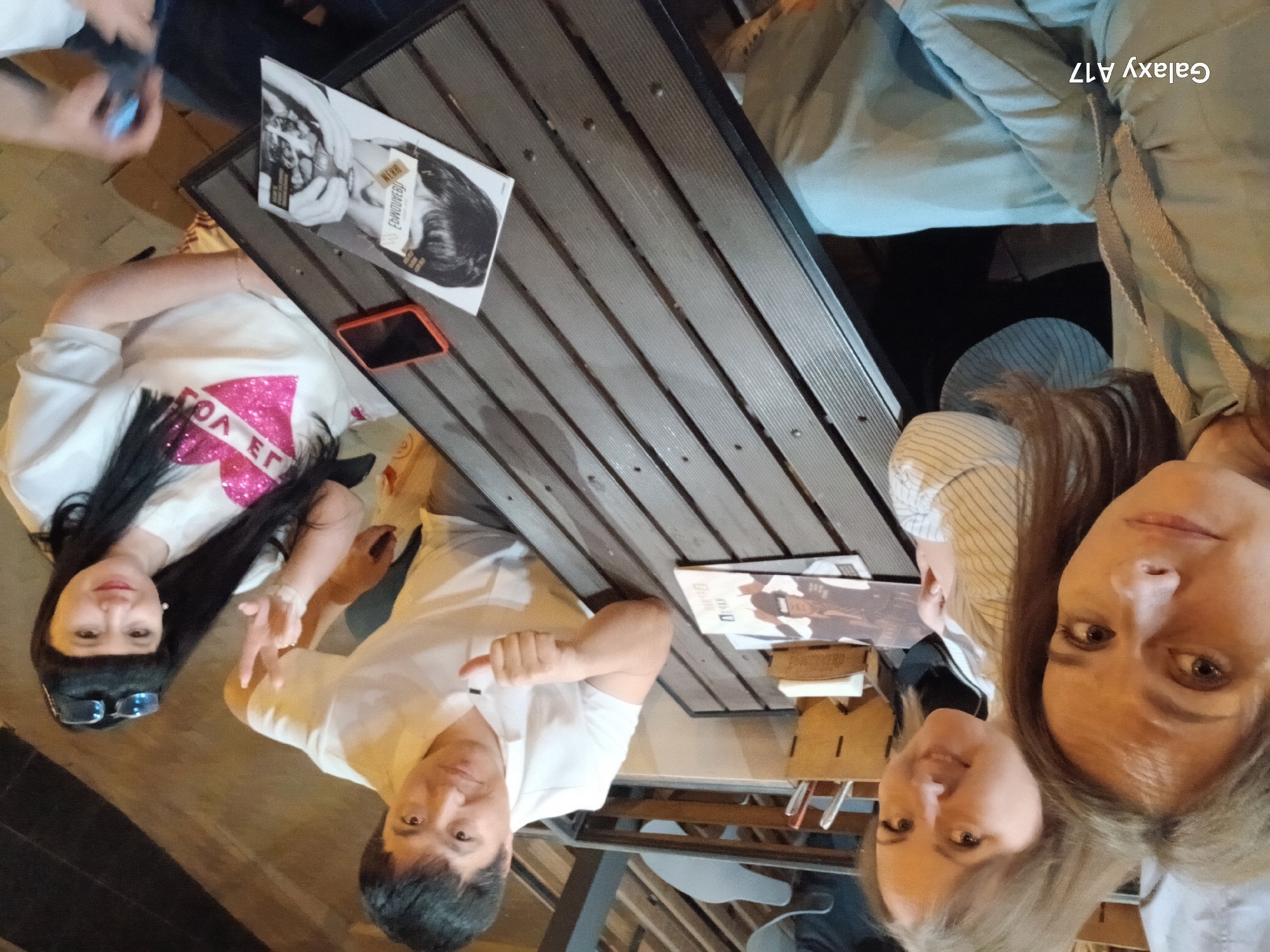 A rotated medium shot shows a group of people sitting at a picnic-style wooden table, with the table oriented diagonally across the frame.
Four people are visible. From left to right: a woman with long black hair wearing a white shirt with a pink heart design, giving the peace sign; a man in a white shirt pointing to the right; and two women at the right of the frame. One woman is wearing a beige blouse with white stripes, and the other is in a light green jacket.
On the table are two magazines. There is also a small red smartphone.
The setting appears to be an outdoor seating area, possibly a restaurant or cafe. There are metal chairs and parts of other tables visible in the background.
The photo's metadata indicates it was taken with a Galaxy A17. Although the photo does not contain information to confirm the location is in Yekaterinburg, Russia, this information was provided separately. The image itself does not visually confirm this.