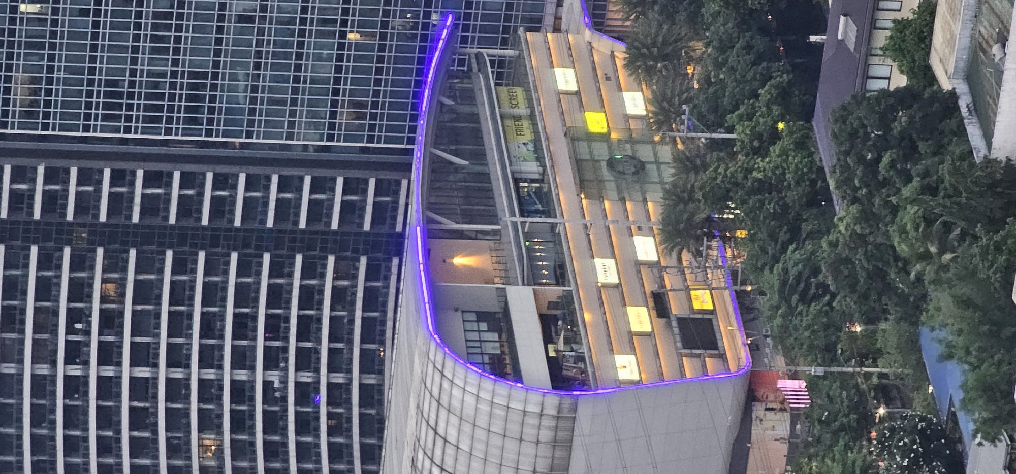 The image shows a cityscape in Makati, Philippines, featuring a modern building with a distinctive curved roof illuminated by purple lights. The building's facade is adorned with various lit signs, suggesting a commercial establishment. Behind this building, taller skyscrapers dominate the skyline, with their windows reflecting the ambient light. Lush green trees partially obscure the lower parts of the structures, indicating a well-treed urban environment. The scene appears to be captured at dusk or during twilight hours, as evidenced by the artificial lighting and the subdued natural light.