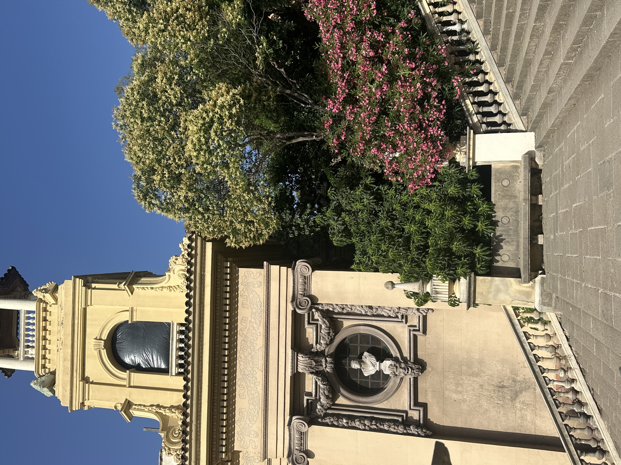 The image shows an exterior view of a building in Barcelona, Spain, likely the Parc del Laberint d'Horta, a historic garden complex. The scene features a tall, light yellow building with classical architectural elements, including columns, decorative carvings, and a balustrade. A prominent arched window on the building is covered with a black tarp.
To the right of the building, lush greenery is visible, including a large tree with pale blossoms and a dense bush with bright pink flowers. A stone staircase with a decorative balustrade ascends from the foreground towards the right side of the frame. In the foreground, there are stone pavers. A stone bench is situated between the building and the staircase. The sky is clear and blue. There are no people visible in the image.