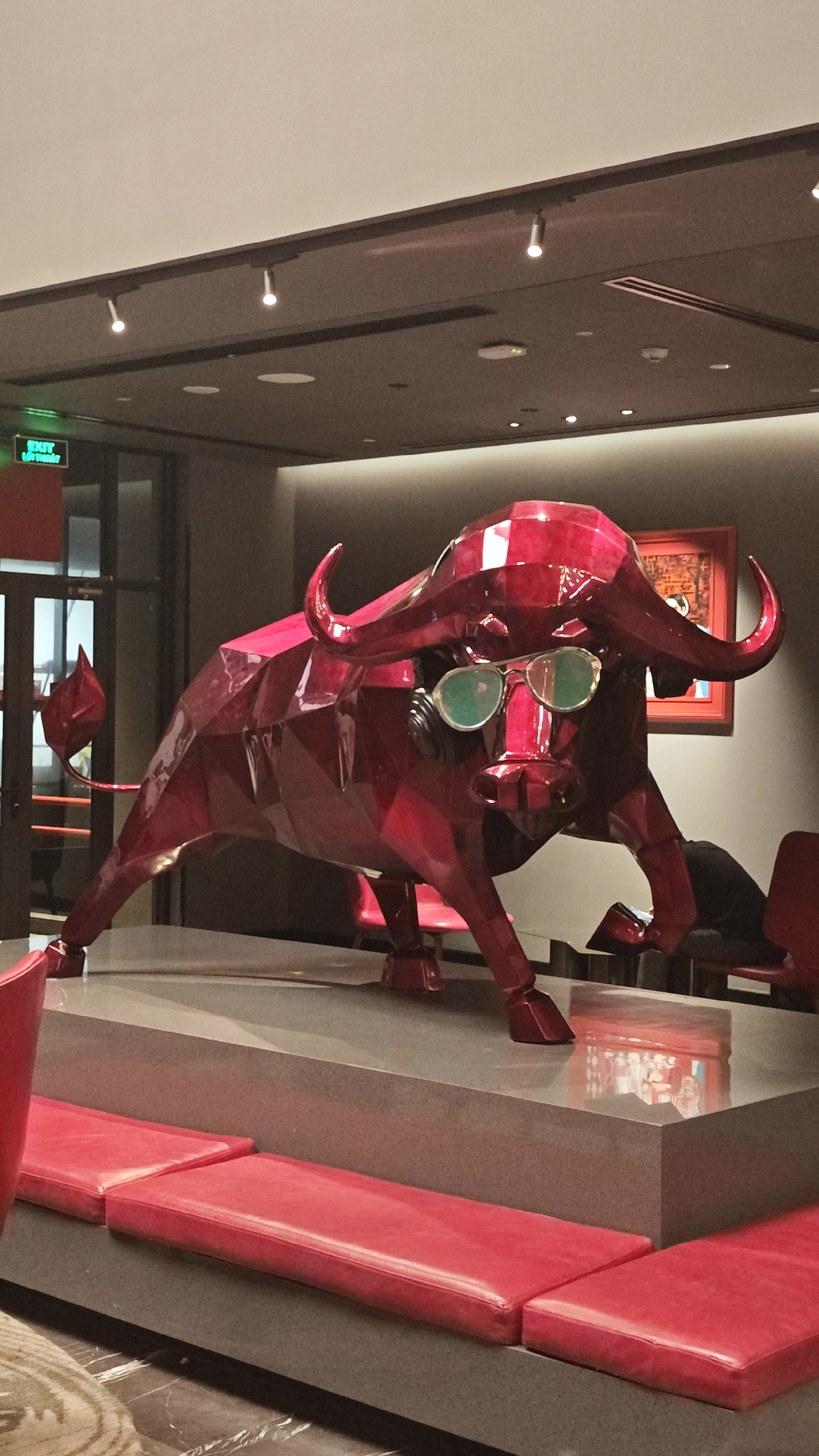 This image presents a modern interior space, likely a hotel lobby or lounge in Đà Nẵng, Vietnam. The central feature is a large, vibrant red sculpture of a bull, rendered in a polygonal, faceted style, resembling a low-poly 3D model or a cut gemstone. The bull is depicted in a powerful, forward-leaning posture, and uniquely, it wears a pair of round, green-tinted sunglasses, adding a whimsical and contemporary touch.
The sculpture stands on a grey, elevated platform. In front of this platform are matching red, upholstered benches, suggesting a waiting or lounge area. The surrounding walls are dark grey, providing a sophisticated backdrop. Overhead, the ceiling is equipped with multiple recessed spotlights and track lighting fixtures that illuminate the space.
To the left of the sculpture, a glass door is visible, and above it, a bright green "EXIT" sign is clearly legible, with the Vietnamese translation "LỐI RA" visible beneath it, confirming the location context. To the right, a framed painting hangs on the wall, featuring warm, perhaps abstract, imagery. A person is partially obscured behind the bull's head, sitting on a red chair. The artificial lighting indicates an indoor setting, likely during the evening or night. The overall scene conveys a stylish and artistic atmosphere.