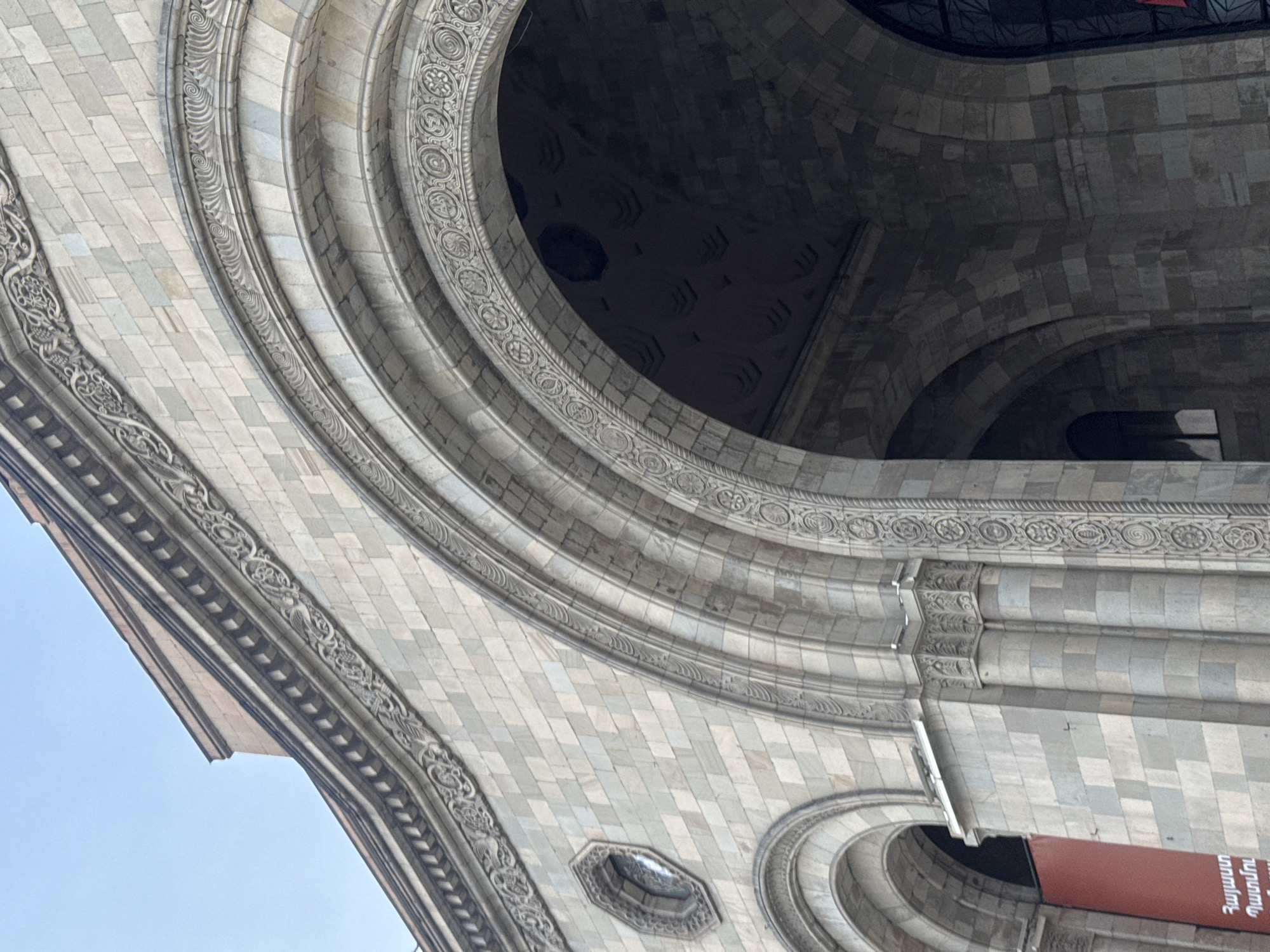 This is an upward-angled photograph showcasing the architectural details of a building in Yerevan, Armenia. The image is dominated by ornate, arched structures made of light-colored stone. The stone is laid in a pattern resembling small bricks or tiles. The arches are adorned with intricate carvings and moldings, which feature repeating geometric and floral designs. Some arches reveal dark, shadowed interiors, hinting at internal spaces. The sky is visible in the lower-left corner.