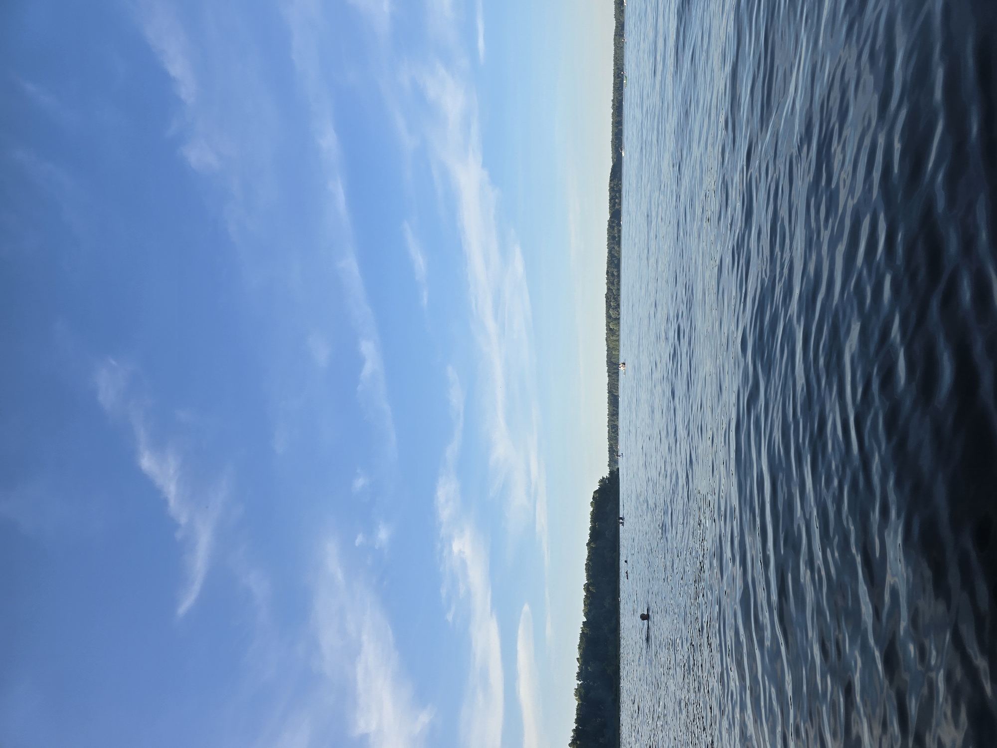 The image depicts a wide, calm body of water, likely a reservoir given the reference to the Kuybyshev Hydroelectric Station. The water is a deep blue, with gentle ripples reflecting the light. In the distance, a lush green forest line stretches across the horizon, with a clear blue sky above, dotted with wispy white clouds. Several people are visible in the water, some swimming and others on paddleboards. A few boats are also present, scattered across the expanse of the water. The overall impression is one of a peaceful, recreational day on the water, possibly at a designated swimming or boating area within or near the settlement of Posyolok Gidrouzla Imeni Kuybysheva, Russia.
