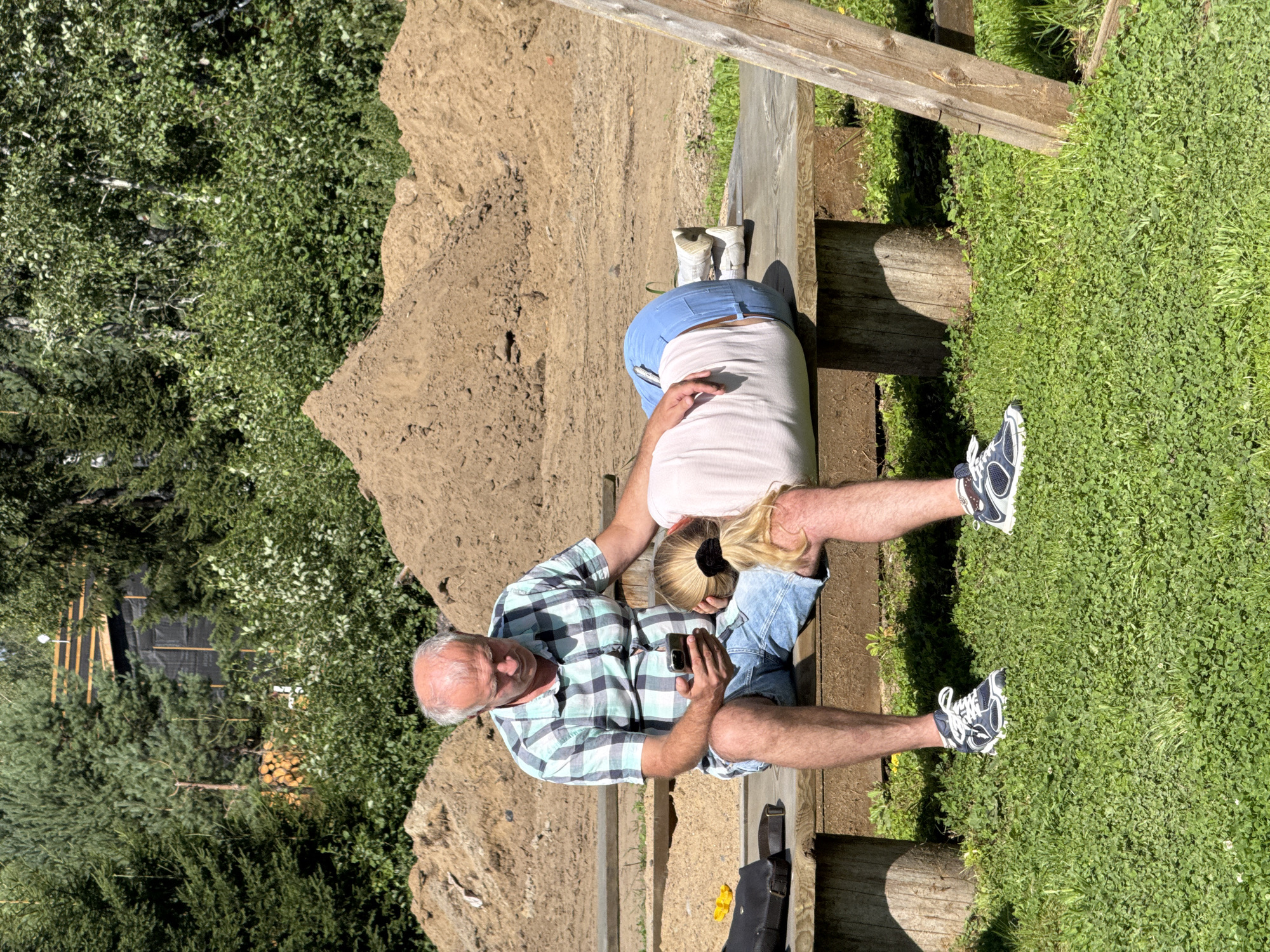 In Khankanmyaki, Russia, a man and a woman are seated on a wooden bench outdoors. The man, balding with a plaid shirt and denim shorts, holds a smartphone. The woman, with blonde hair tied back, rests her head on his lap, wearing a light-colored t-shirt and blue jeans. They are positioned in front of large piles of dirt, with trees and foliage visible in the background under a bright sky.