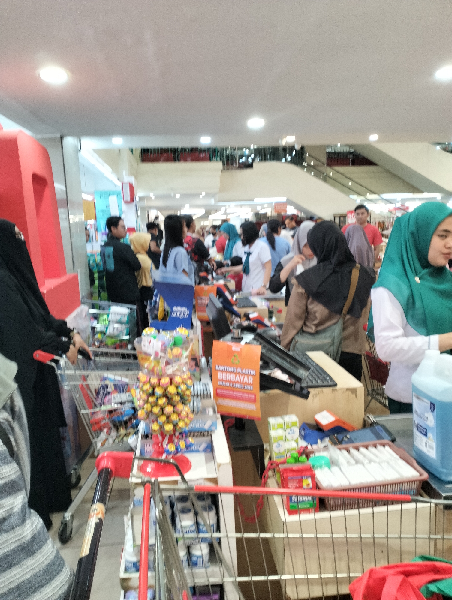 The image captures an indoor scene, likely a supermarket checkout area in Ungaran, Indonesia. A red, partially visible signage with large white lettering stands on the left. In the foreground, the handle and part of a shopping cart are visible, with a basket filled with colorful candies on display. Another shopping cart is situated behind it. Multiple individuals are present, some appearing to be customers and others possibly store employees at the checkouts. One person in the foreground to the left is wearing a black niqab and black clothing, holding the shopping cart. Further back, several people are gathered around checkout counters. A person wearing a green hijab is standing at one of the counters, interacting with a cash register. The environment appears to be brightly lit, with white ceiling panels and recessed lighting. Escalators are visible in the background, suggesting a multi-level store. Products such as drinks and cleaning supplies are visible on shelves and at the checkout. The overall impression is a busy commercial setting during operating hours.