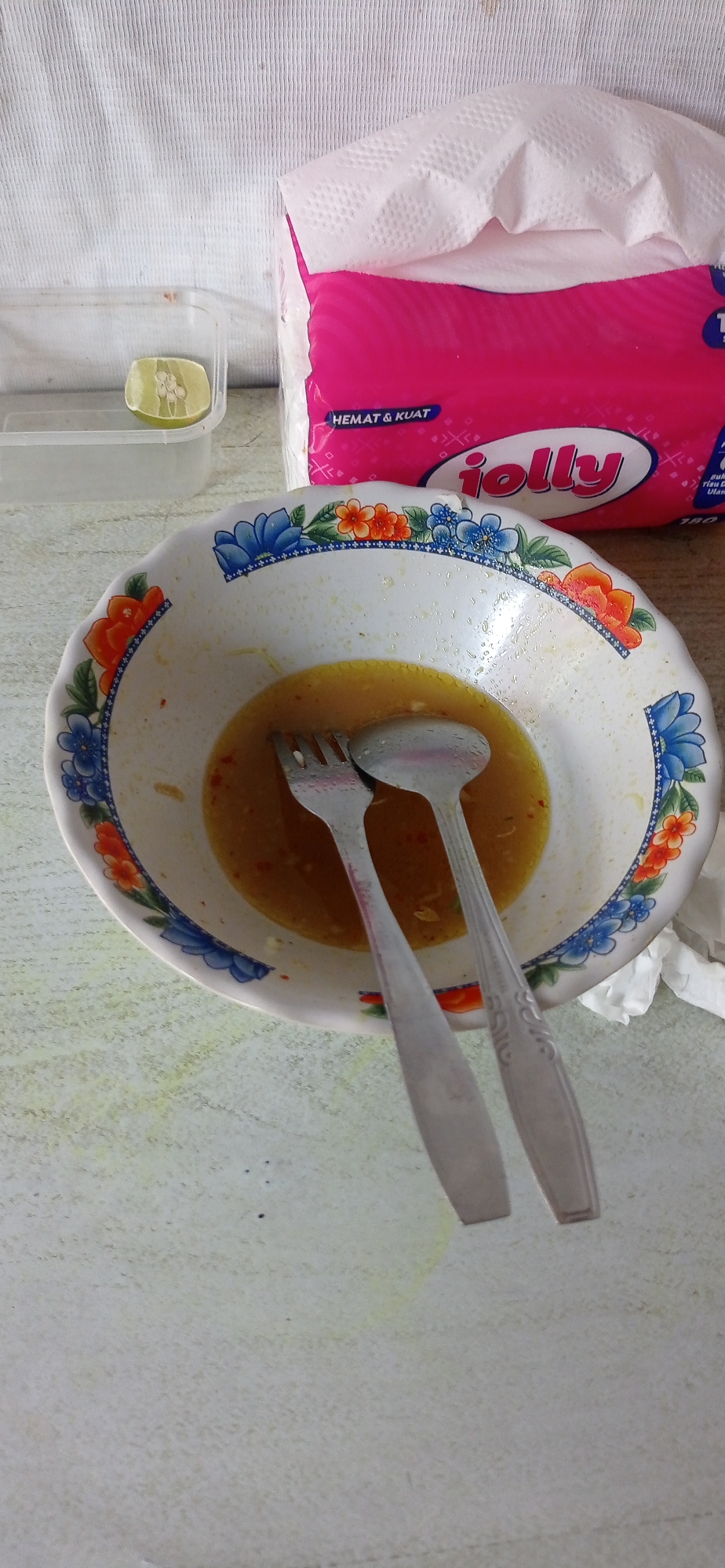 The image depicts a close-up, high-angle shot of a partially consumed meal in a bowl. The bowl, which appears to be ceramic with a floral rim design, contains a yellow liquid, possibly soup or broth, with visible remnants of food particles. A fork and a spoon are resting inside the bowl, angled against the sides. To the left of the bowl, a clear plastic container holds a slice of lime. In the background, a stack of pink and white paper napkins is partially visible, featuring the "jolly" logo. The overall setting appears to be an indoor surface, possibly a table, with a textured, light-colored finish. The environment suggests a casual dining scenario. The location is identified as Jumputrejo, Indonesia.