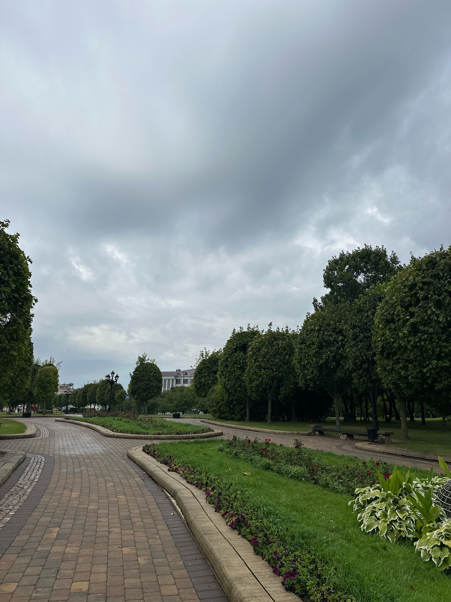 A paved pathway winds through a city park on an overcast day. The pathway is constructed from reddish-brown and grey pavers. On the right side of the pathway, there are green lawns with flowerbeds containing small purple and red flowers, as well as larger variegated hosta plants. Beyond the lawns, rows of meticulously trimmed, rounded trees line the park. In the distance, a large, multi-story building with white walls and dark windows is visible. A lamppost stands amidst the trees. A few benches are scattered throughout the park. The sky is heavily clouded with grey and white tones. The image appears to be taken from a low angle, looking towards the park's interior. This scene is consistent with the environment of Gorky Park in Minsk, Belarus.