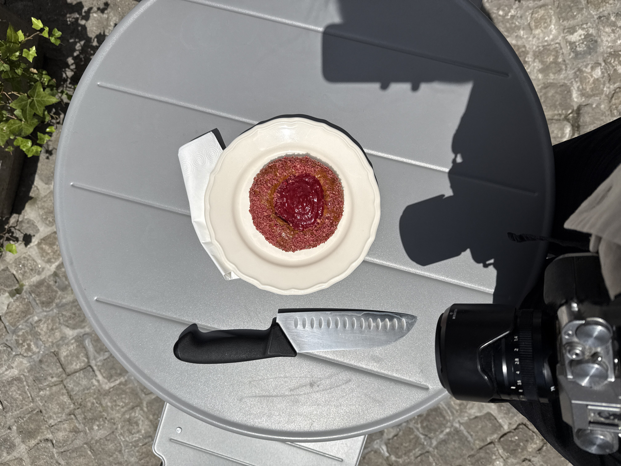 An overhead shot captures a dessert on a round, gray table outdoors, possibly on a patio. To the left of the dessert is a silver knife with a black handle. The dessert, a small pastry with a pink crumbly exterior and a dollop of red jam in the center, sits on a white plate with a ruffled edge. A white napkin is folded beneath the plate. In the foreground, a camera with a black lens hood is visible, partially obscuring the bottom of the frame. The background shows a cobbled stone surface and some green foliage. The scene is bathed in bright sunlight, casting distinct shadows. The image does not provide any specific details or indicators to pinpoint its location within the Vracar Urban Municipality, Serbia.