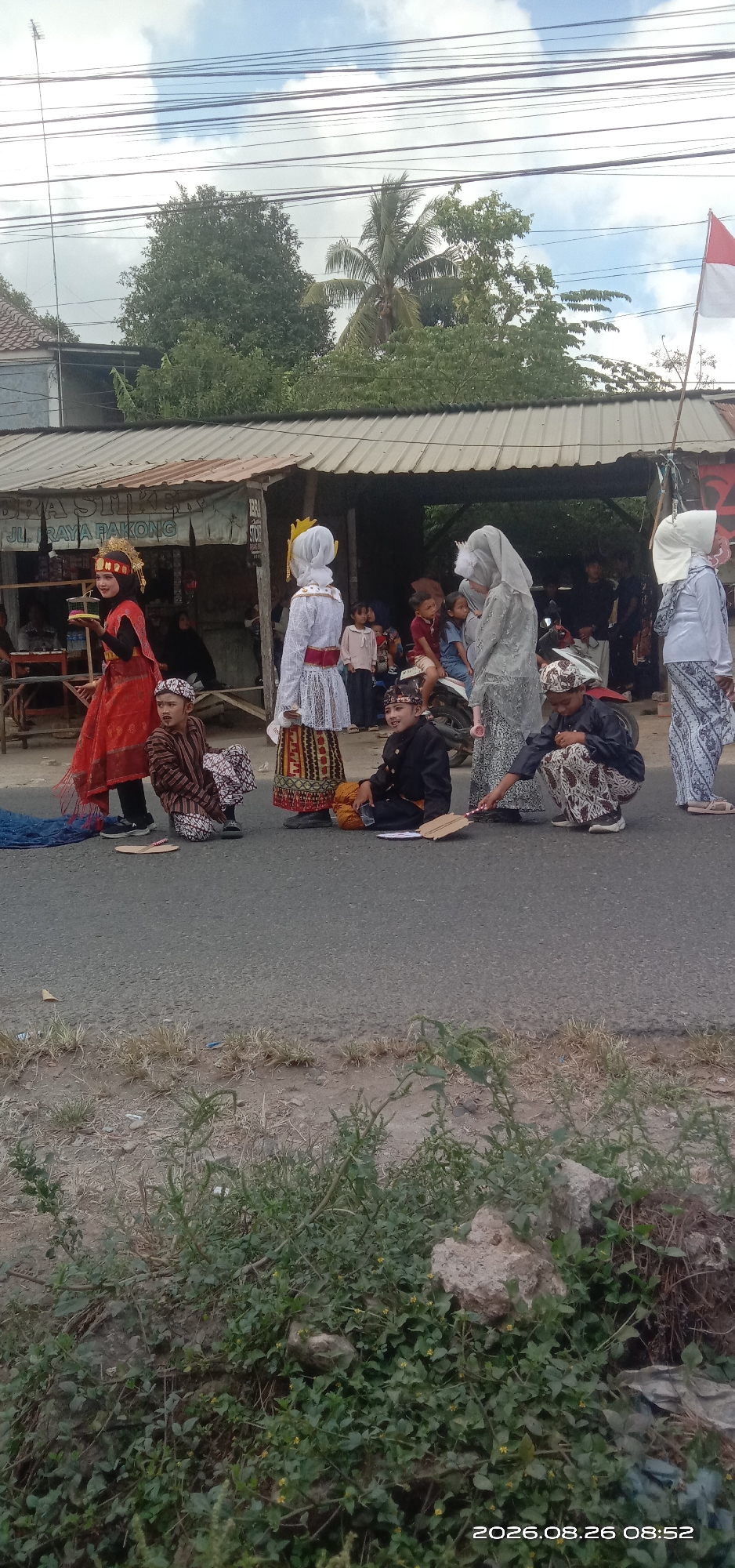 In Pakong, Indonesia, a group of children and young people in traditional attire are gathered on the side of a paved road. A building with a corrugated metal roof and visible signage that reads "JL. RAYA PAKONG" serves as a backdrop. Some individuals are seated on the ground, while others stand. One person in a red traditional dress holds a yellow object. Two individuals in black traditional outfits are seated on the ground, one holding a fan-like object. Another person in a light-colored traditional outfit with a headscarf stands and appears to be interacting with a person in a similar outfit to their right. A red and white flag is visible in the upper right corner of the image. The environment suggests an outdoor event or performance taking place near a commercial area.
