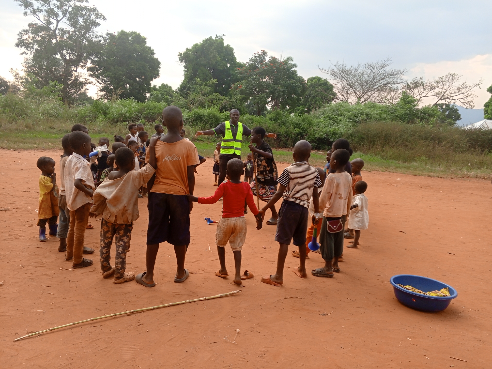 In Ngalula, Democratic Republic of the Congo, a group of children stand in a circle on a dirt ground. An adult male in a gray shirt and a bright yellow safety vest stands among the children, his arms outstretched. A blue tub sits on the ground near the edge of the circle. A long, thin stick lies on the ground near the children's feet. The background includes trees and vegetation under a cloudy sky.