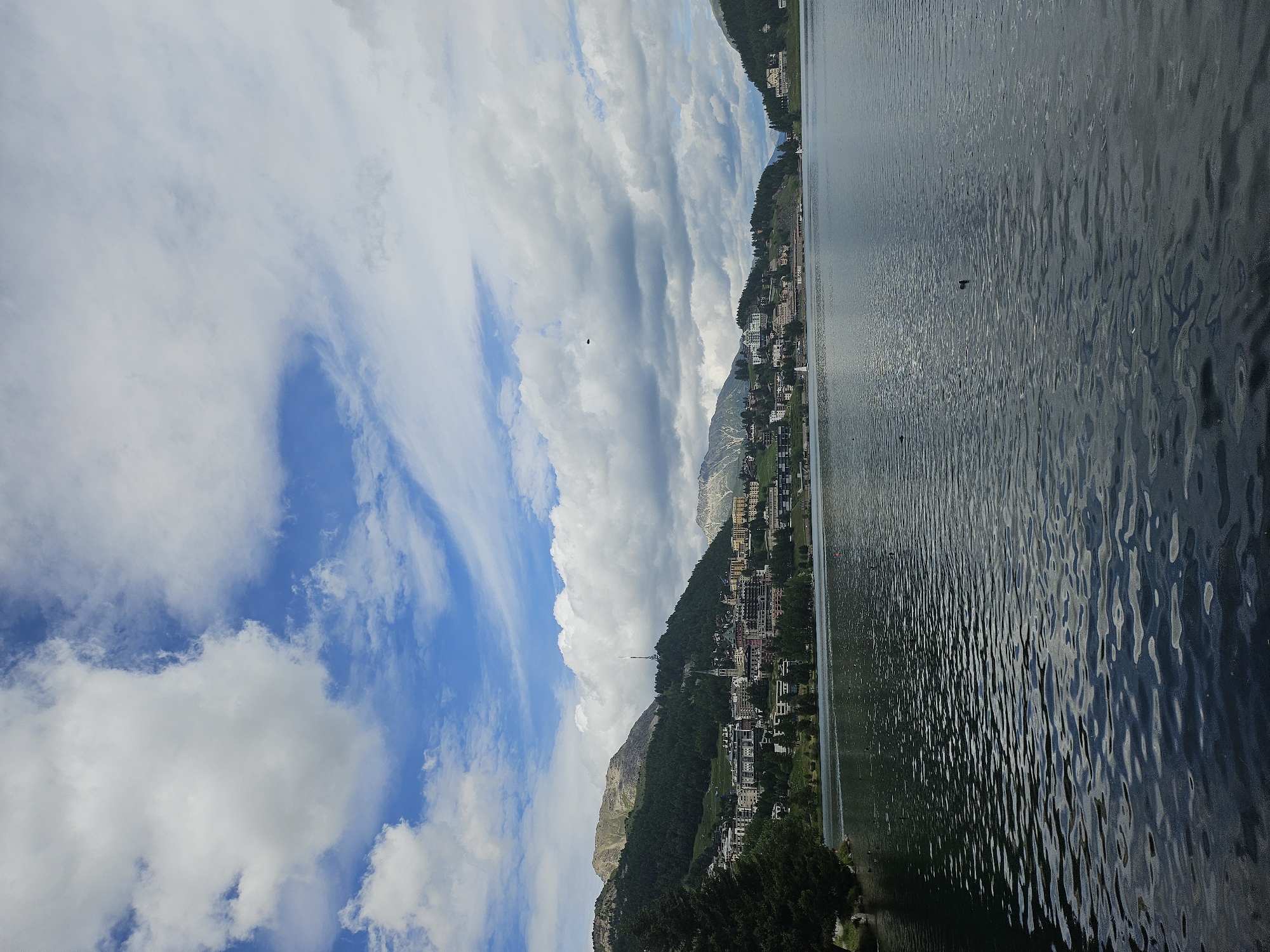 This is a vertical, wide-angle shot of a scenic lake with a town nestled on a hillside in St. Moritz, Switzerland. The sky is a vibrant blue with dramatic, wispy white clouds that create a sense of movement and depth. The sun is shining, casting reflections on the rippling surface of the dark, calm water.
On the left side of the image, a lush green hillside is covered with trees and dotted with buildings that make up the town of St. Moritz. The architecture appears to be a mix of traditional and modern styles, with many buildings featuring balconies and pitched roofs. Some of these buildings are hotels or apartments, indicating a tourist destination. The town stretches along the hillside, creating a picturesque urban landscape against the natural backdrop of the mountains.
On the right side of the image, the lake dominates the frame. Its surface is textured with small waves, reflecting the sky and the surrounding landscape. A few dark shapes are visible on the water, possibly birds or small boats. The overall impression is one of tranquility and natural beauty, with the town adding a touch of human settlement to the serene environment. The time of day appears to be daytime, likely late morning or early afternoon, given the brightness of the sunlight and the clear sky with scattered clouds. The weather is pleasant and fair.