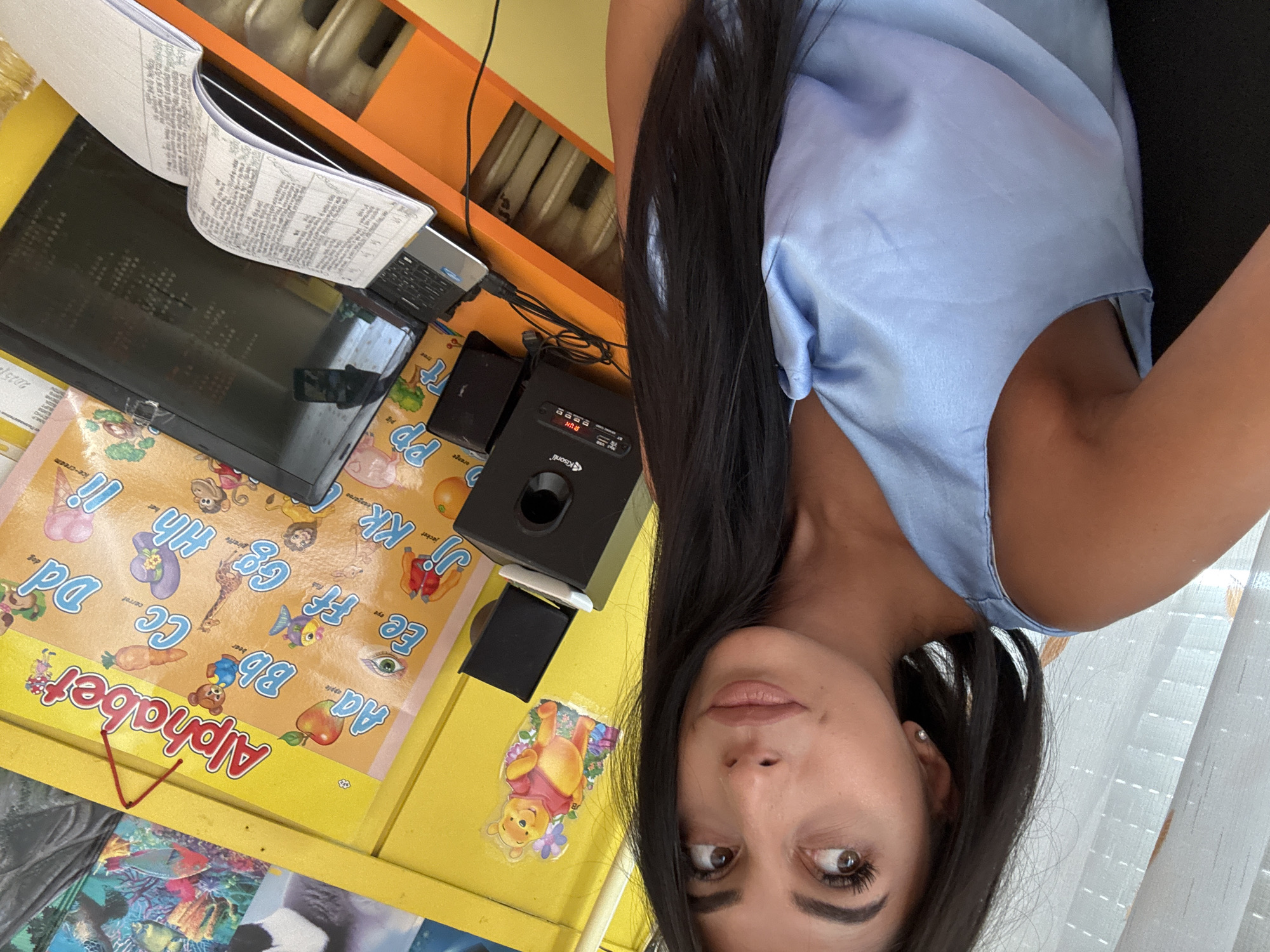 A medium shot shows a woman in a light blue tank top looking to her right with a slight smile. She has dark hair and fair skin. Behind her, an alphabet poster hangs on a yellow wall. A small black speaker sits on a desk in front of the poster, with a laptop and an open notebook to its right. The environment appears to be indoors, possibly a classroom or office space. The presence of an alphabet poster suggests a learning or educational setting. The image does not provide any specific clues to the location in the city of Plovdiv, Bulgaria.