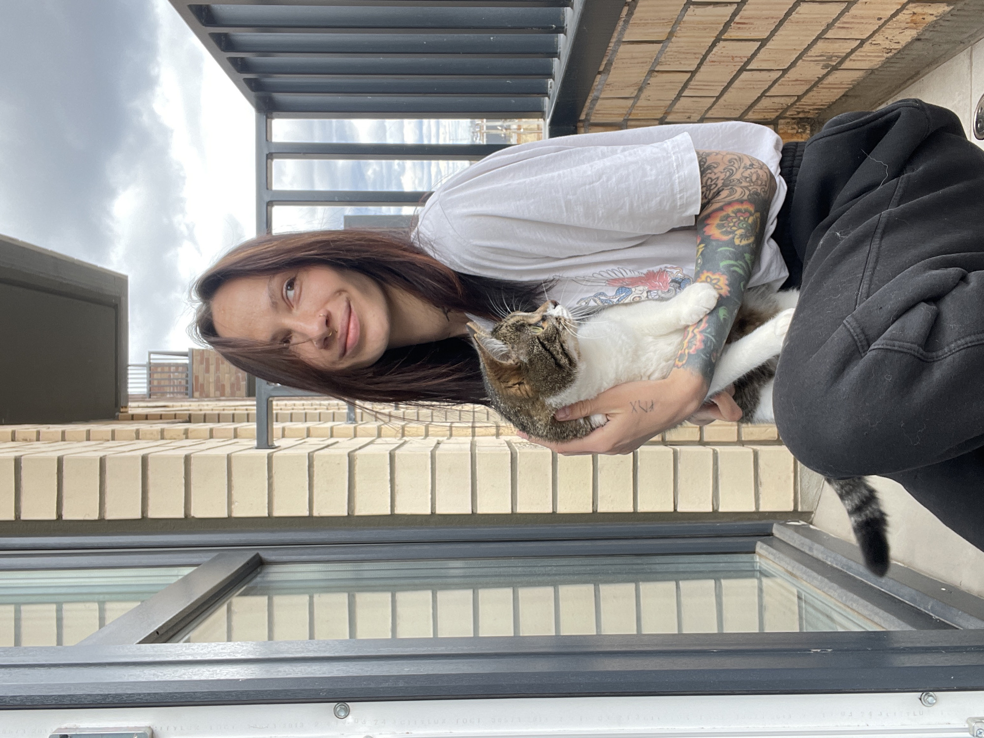 The image, when correctly oriented, depicts a young woman affectionately holding a tabby and white cat on what appears to be a balcony or outdoor terrace. The setting is under an overcast sky, suggesting a cloudy day.
The woman has long, dark brown hair and a gentle smile, with a nose piercing. She is wearing a white t-shirt and black sweatpants. Both of her arms are extensively tattooed; the visible left arm features a colorful sleeve with floral motifs, while the right arm displays intricate blackwork patterns. On the back of her left hand, a small tattoo resembling "XVY" or "XXV" (Roman numerals) is visible.
She is cradling a fluffy cat, which is predominantly white with brown and black tabby markings on its head, back, and tail. The cat looks upward, seemingly content in the woman's arms.
The background shows a building's exterior, likely part of the same residential complex. A light-colored brick wall forms part of the terrace, above which a dark metal railing system is visible. The sky is filled with grey clouds. Below the subjects, a large window or glass panel with a dark frame is prominent, reflecting the outdoor environment. Faint text is visible on the bottom frame of the window, including what appears to be "ГОСТ 30674-2013", a Russian standard for window blocks.
The image captures a quiet, affectionate moment between a pet owner and her cat in an outdoor urban or suburban setting, likely in Pervomaysky, Russia, as indicated by the location context and visible GOST standard on the window frame.